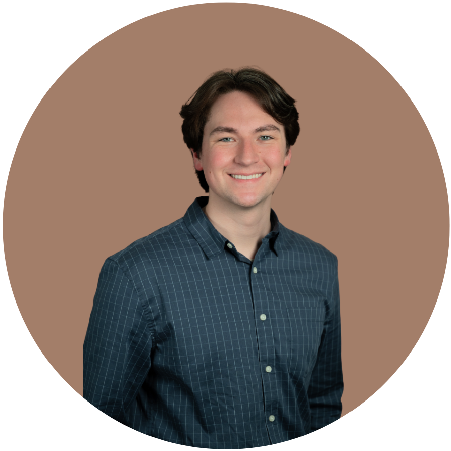 A young man with brown hair and blue eyes, smiling, wearing a blue checkered button-down shirt, standing in front of a beige background.