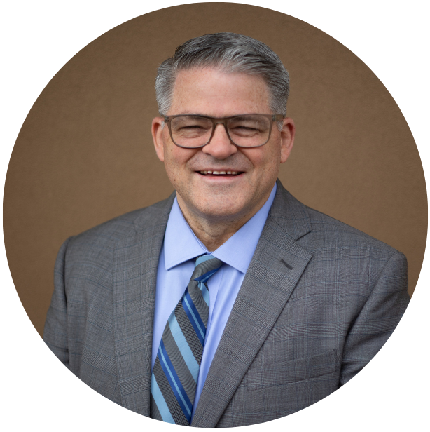 Portrait of a middle-aged man wearing glasses, a gray checkered suit, light blue shirt, and blue striped tie, smiling against a brown background.