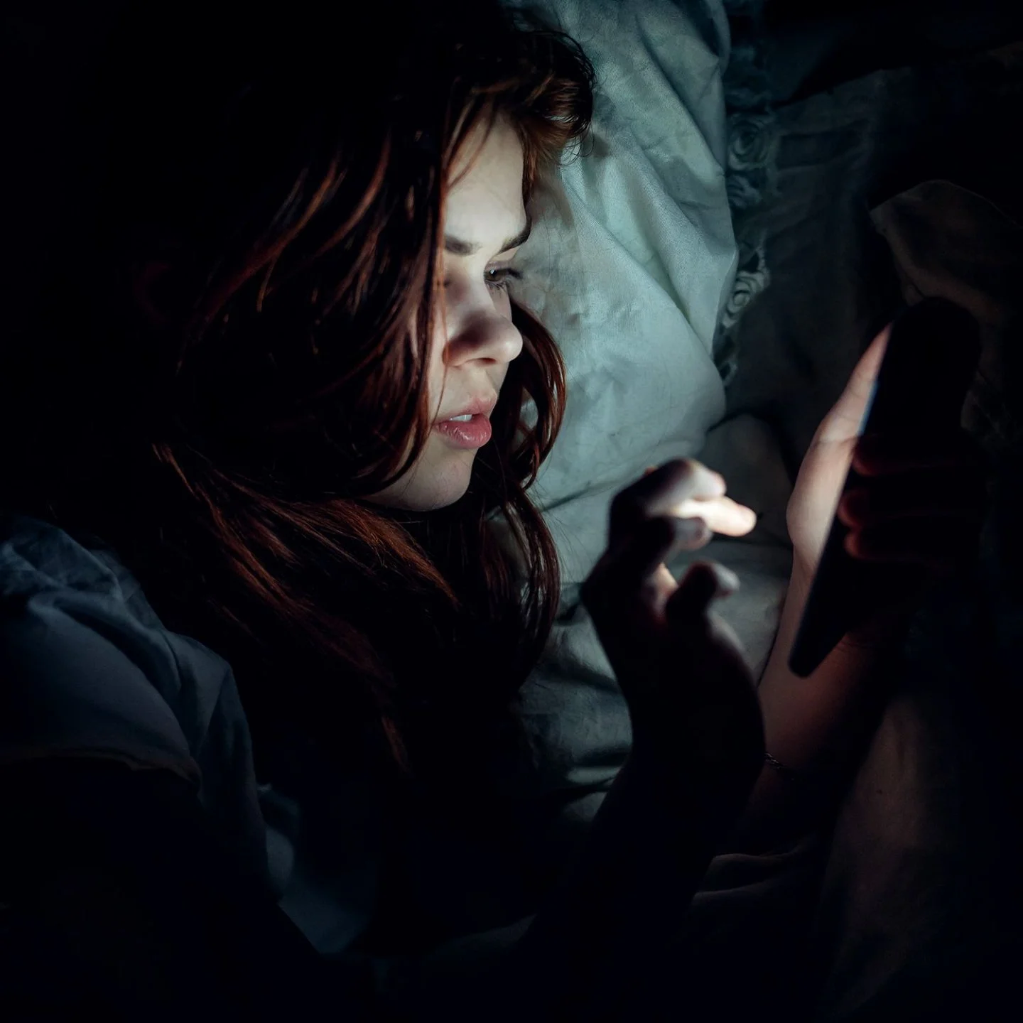 A woman with red hair lying on a bed, illuminated by her phone screen, with pillows and blankets around her.