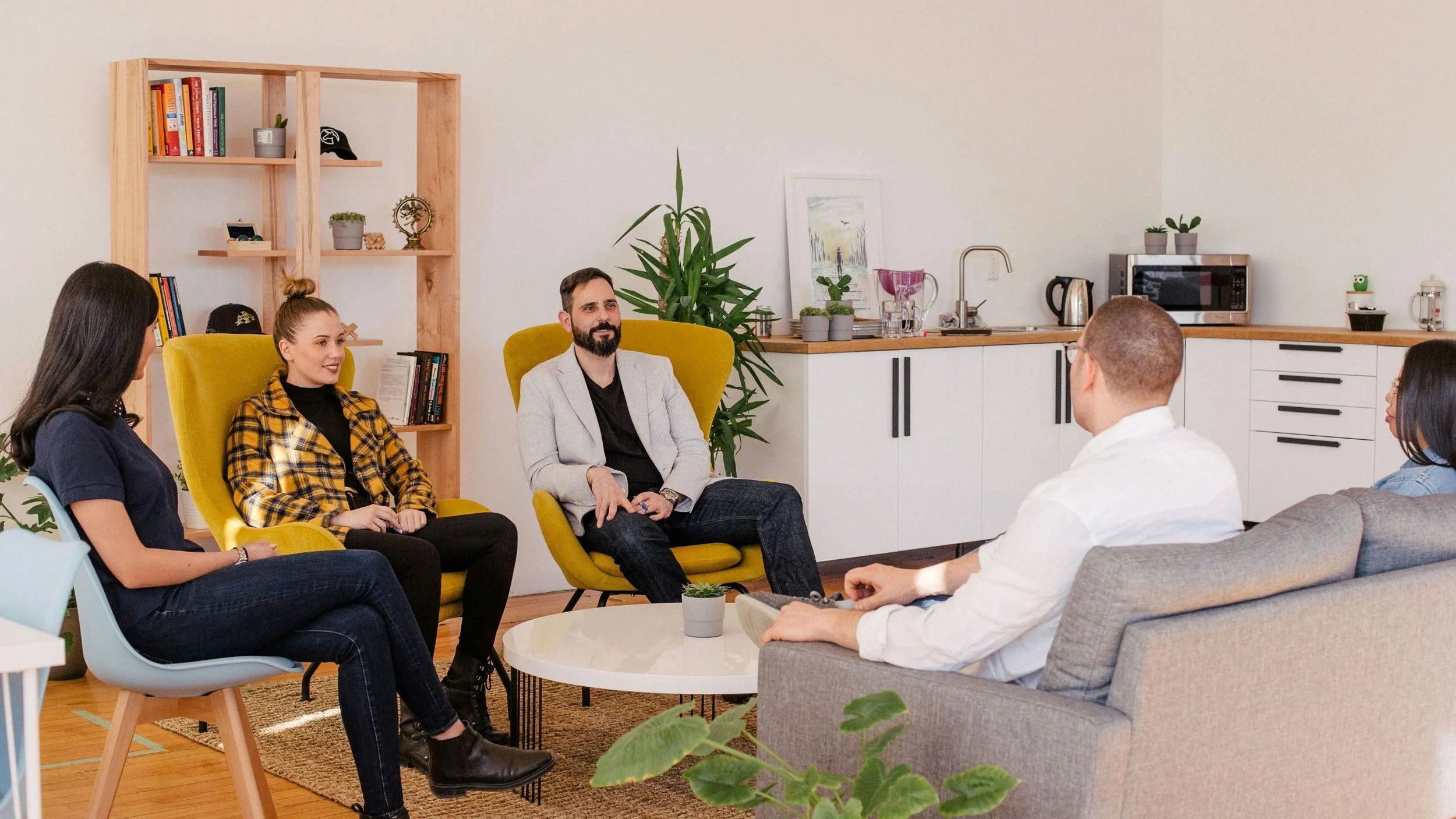A group of five people having a discussion in a cozy, modern kitchen and living area, with plants and books on shelves.