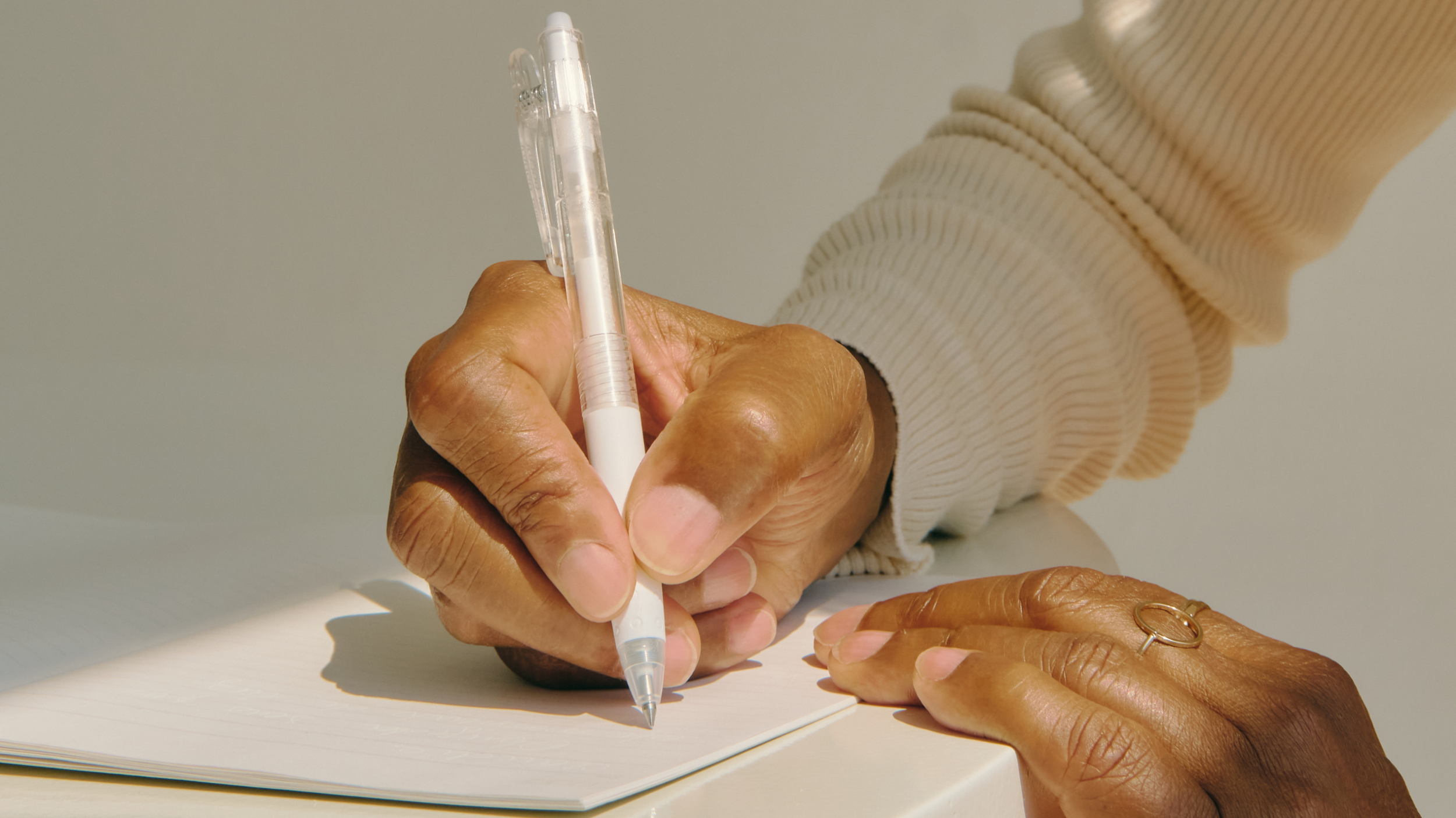 Close-up of a hand writing in a notebook with a white pen, with another hand resting on the table, wearing a gold ring.