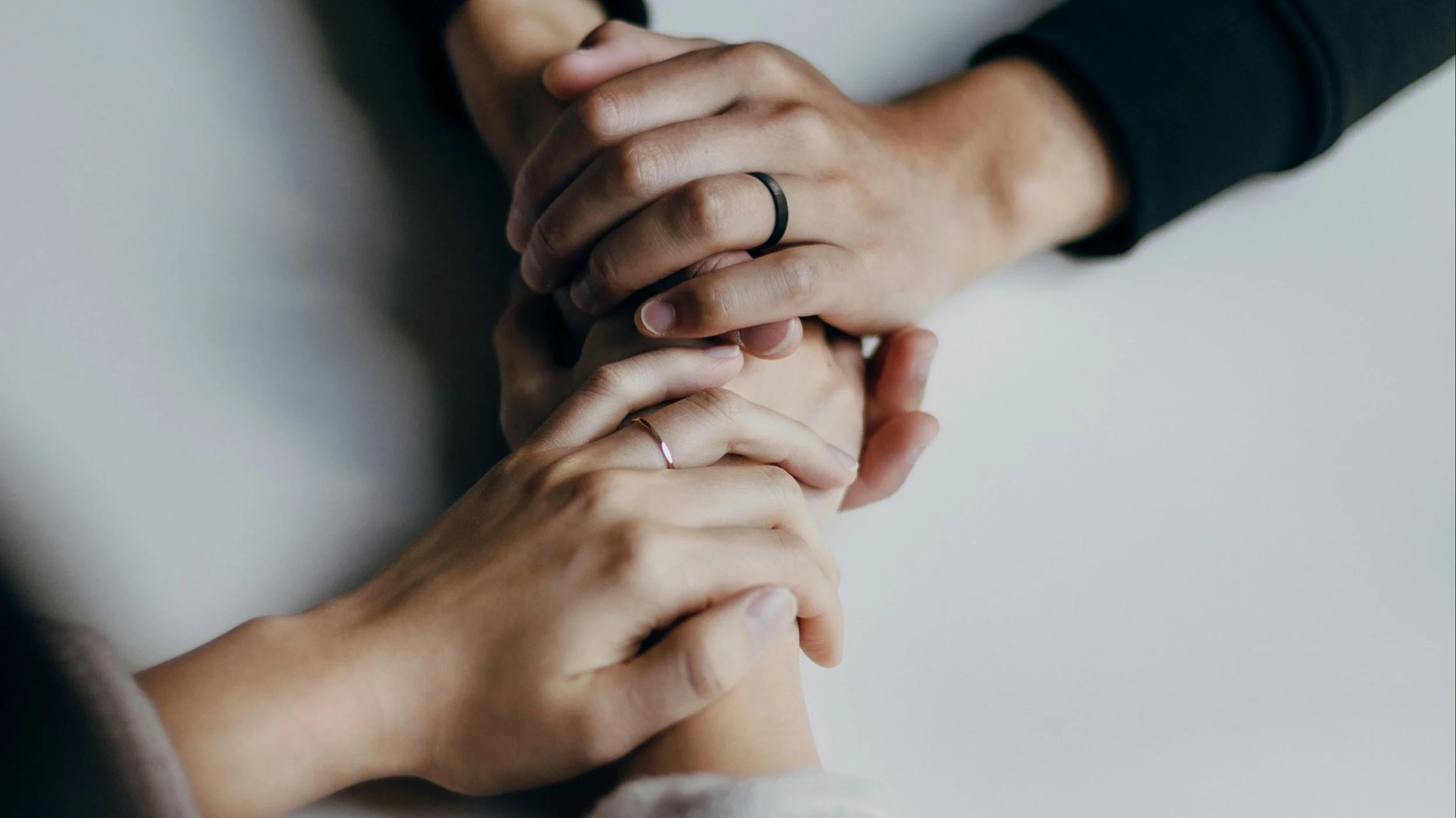 Multiple hands gently holding each other, symbolizing support and connection, with some hands wearing rings, against a plain light background.