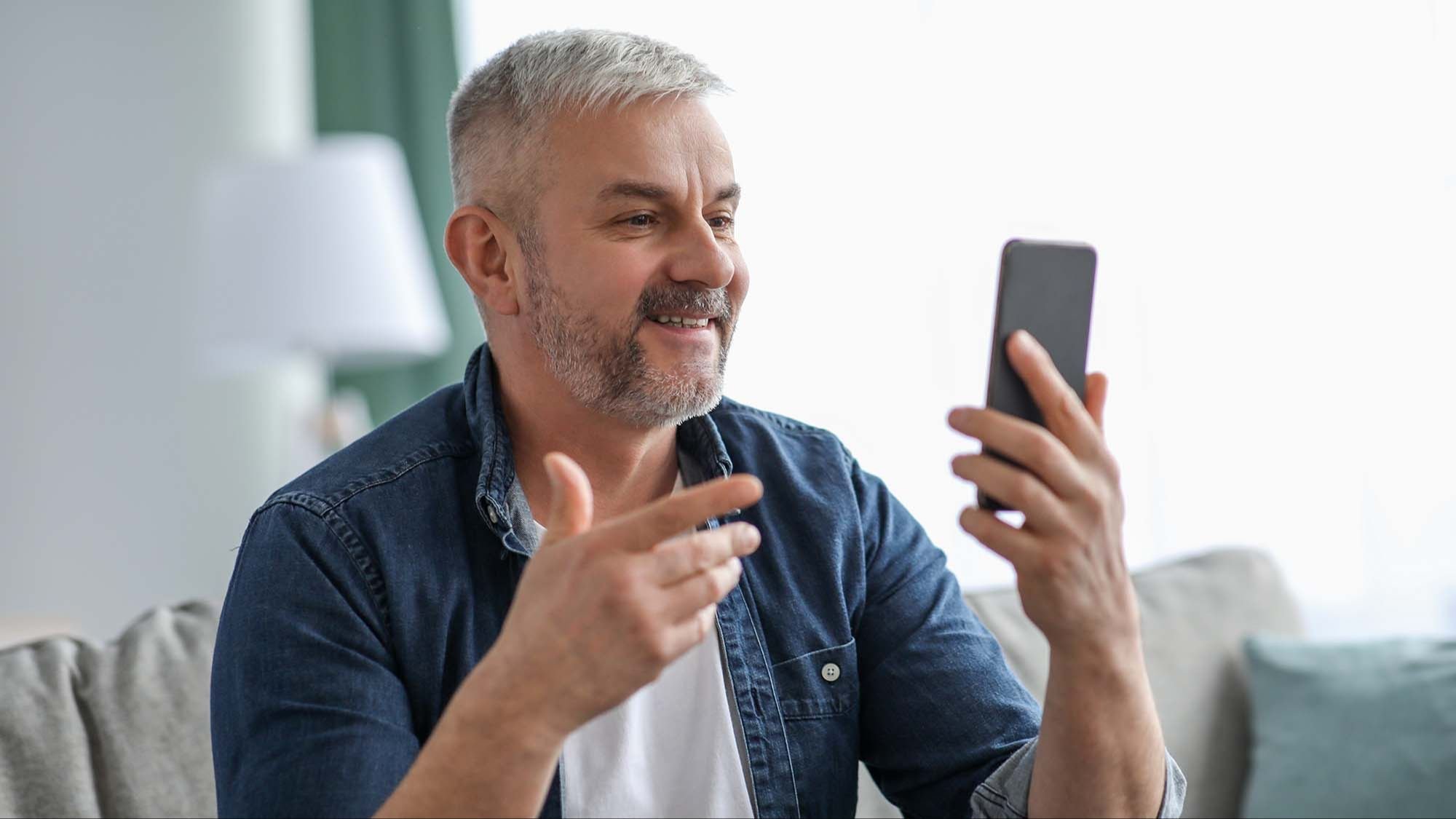 A smiling middle-aged man with gray hair and a beard looking at his smartphone while sitting on a sofa in a bright living room.
