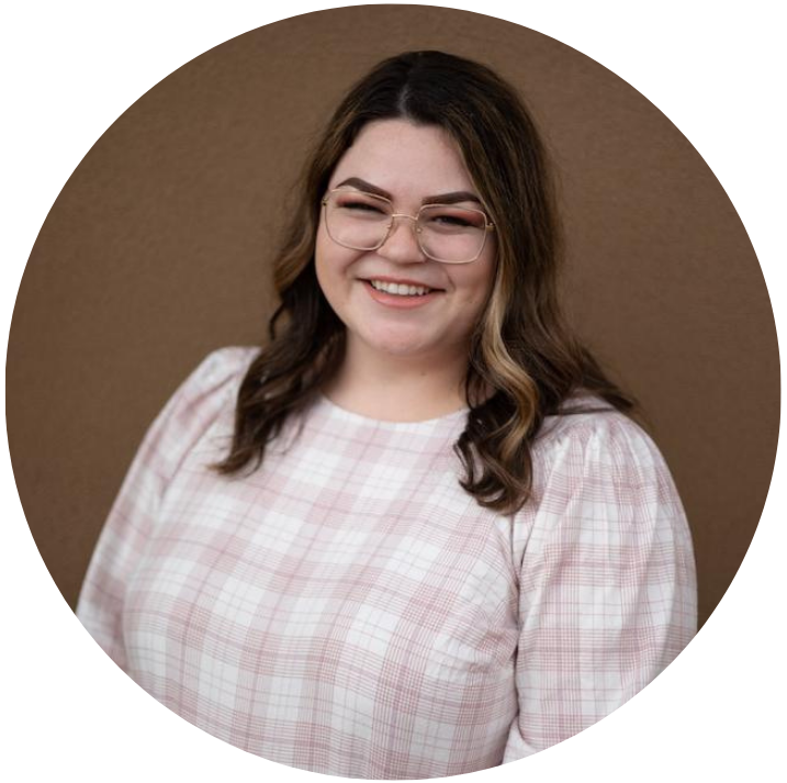 A young woman smiling and wearing glasses, with curly brown hair, in front of a brown background.