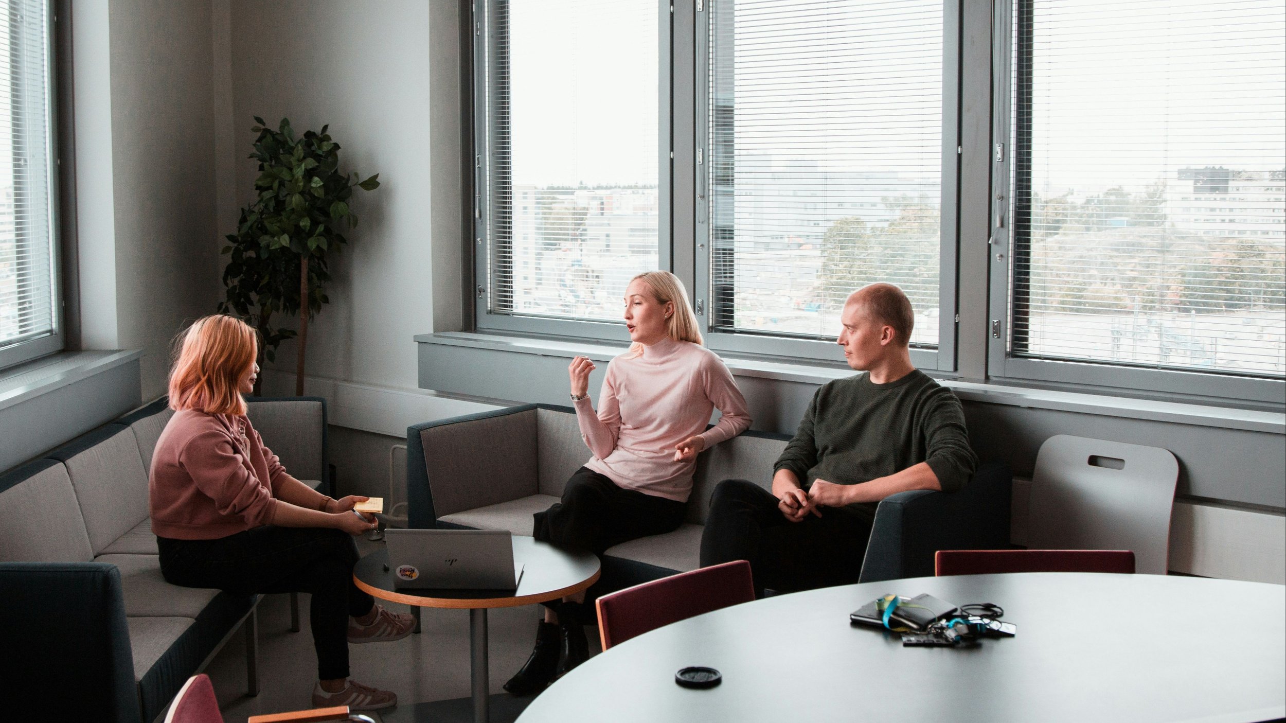 Three people having a conversation in a modern office lounge area with large windows and city view. One woman with red hair sits on the left, another woman with blonde hair in the middle, and a man with short hair on the right. A laptop, some papers, and a few personal items are on the tables.