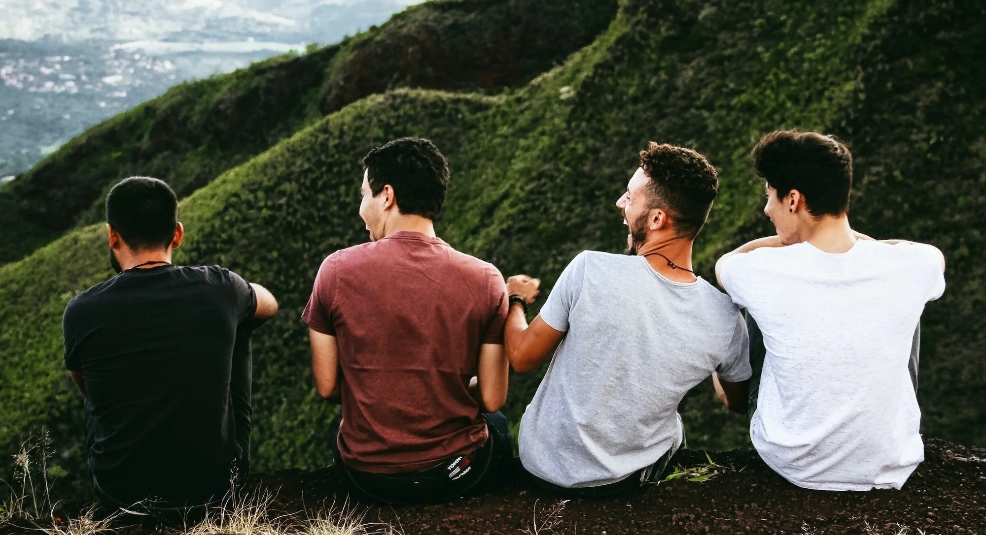 Four young men sitting on a grassy hillside overlooking a lush green valley and distant cityscape, enjoying the outdoors.