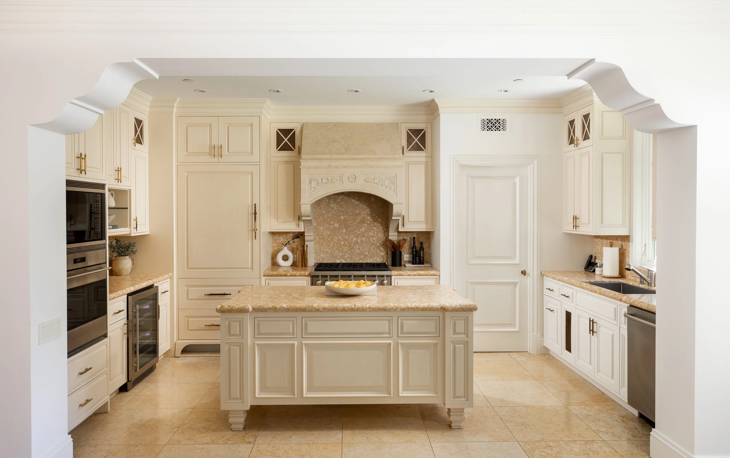 A cream-colored kitchen with white cabinetry, a central island with a beige marble countertop, and a decorative fireplace. The kitchen features built-in appliances, a sink, and various kitchen accessories, with beige tiled flooring and crown molding 