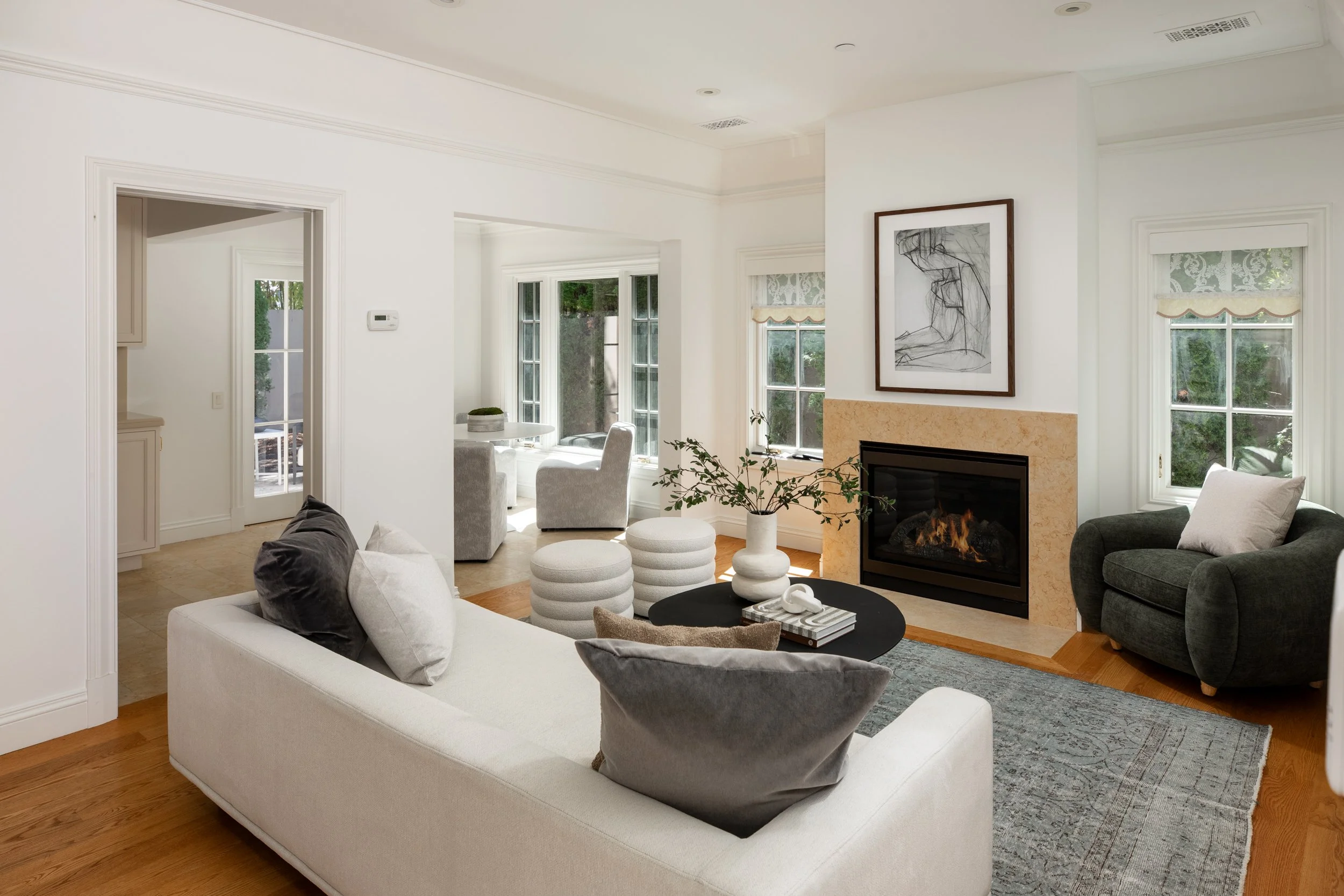 A bright living room with white walls, wooden flooring, and multiple large windows with patterned blinds. It is furnished with a white sofa, dark sofa, several round ottomans, and a black table with books and decorative objects. There is a fireplace 