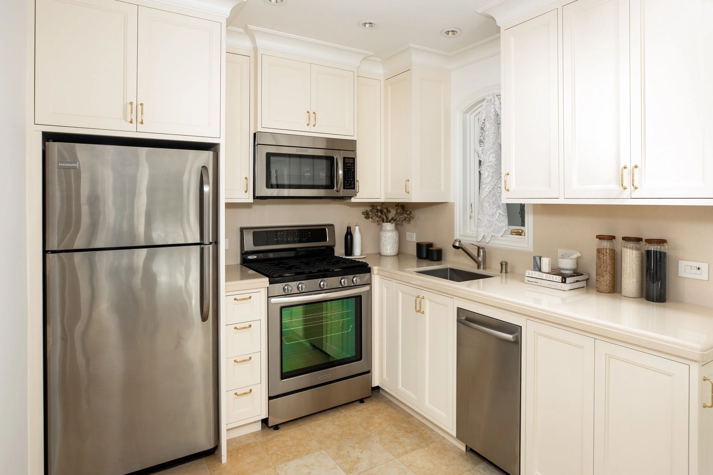 A modern kitchen with white cabinets, stainless steel appliances including a refrigerator, microwave, and oven, a small window with lace curtains, and a countertop with various jars and decorative items.