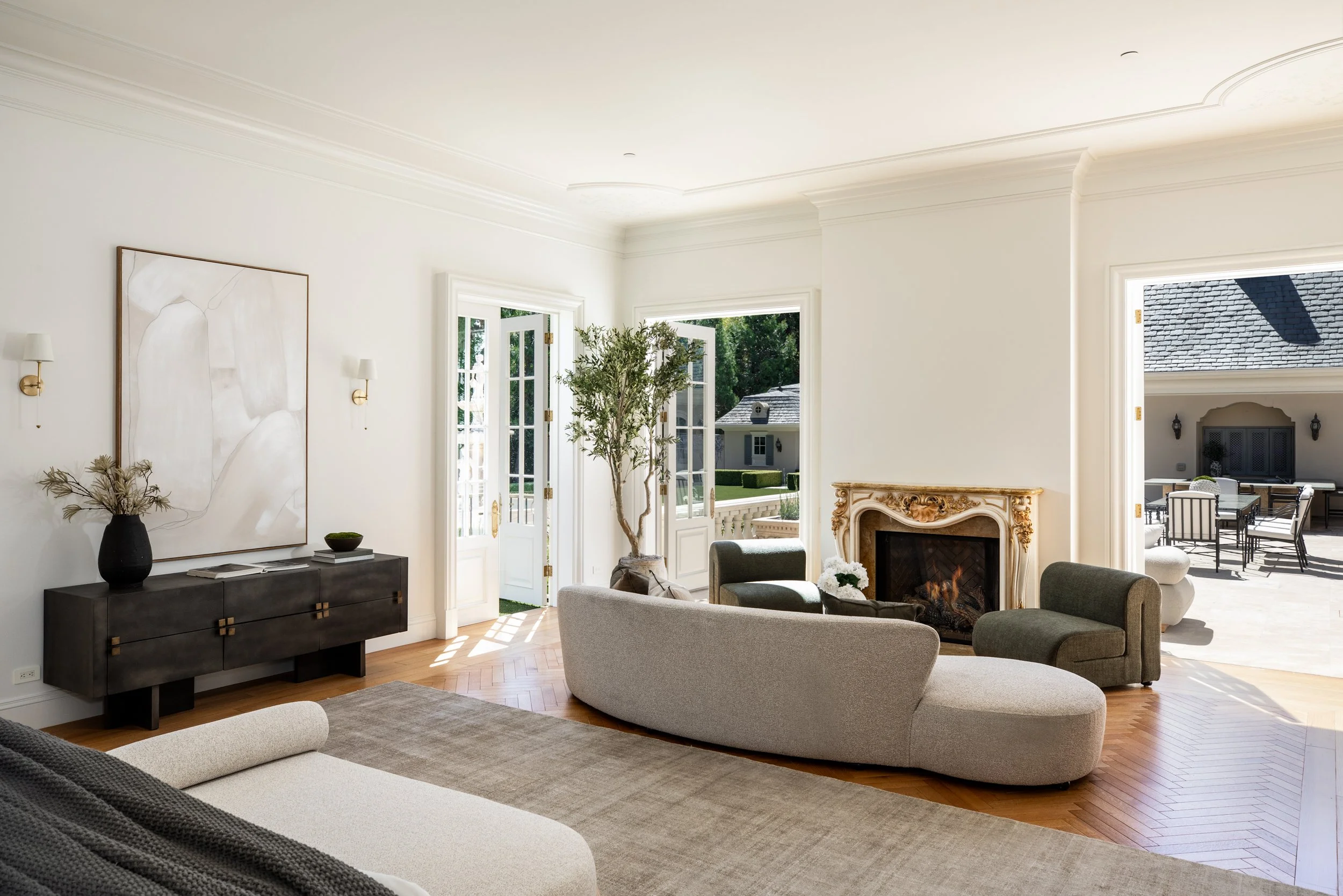 Bright living room with white walls, multiple windows, and comfortable chairs near a fireplace, with a view of an outdoor patio and garden.