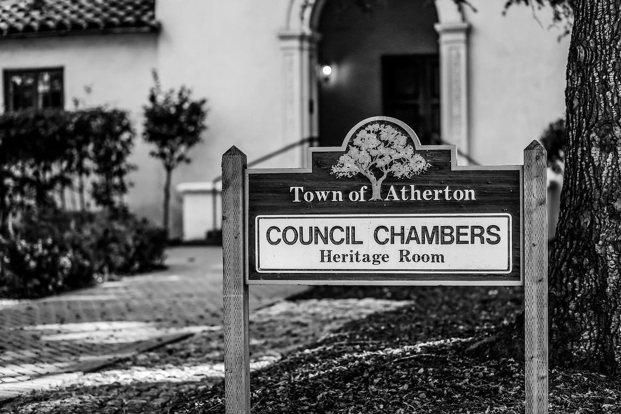 A sign in front of a building indicating it is the Council Chambers Heritage Room in the town of Atherton. The sign has a tree graphic and is situated on a sidewalk near a tree trunk, with some landscaping and the building entrance visible in the bac