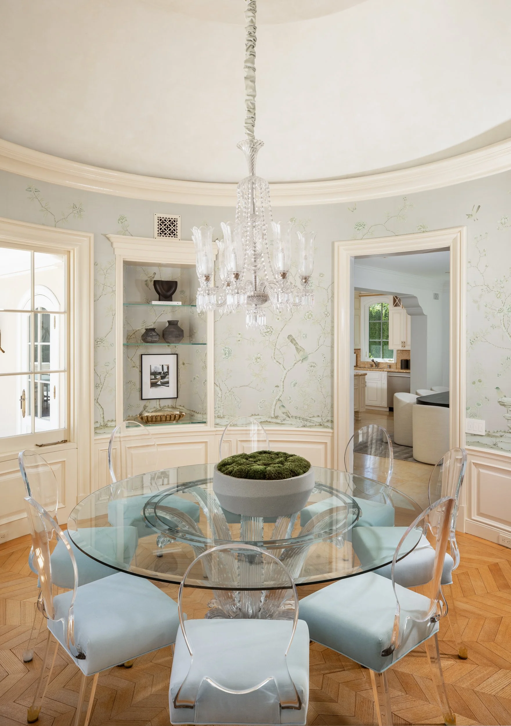 A dining room with a glass top round table surrounded by white cushioned chairs. The table has a decorative bowl with moss in the center. A crystal chandelier hangs from the ceiling. There are windows and doorways leading to other rooms, with built-i