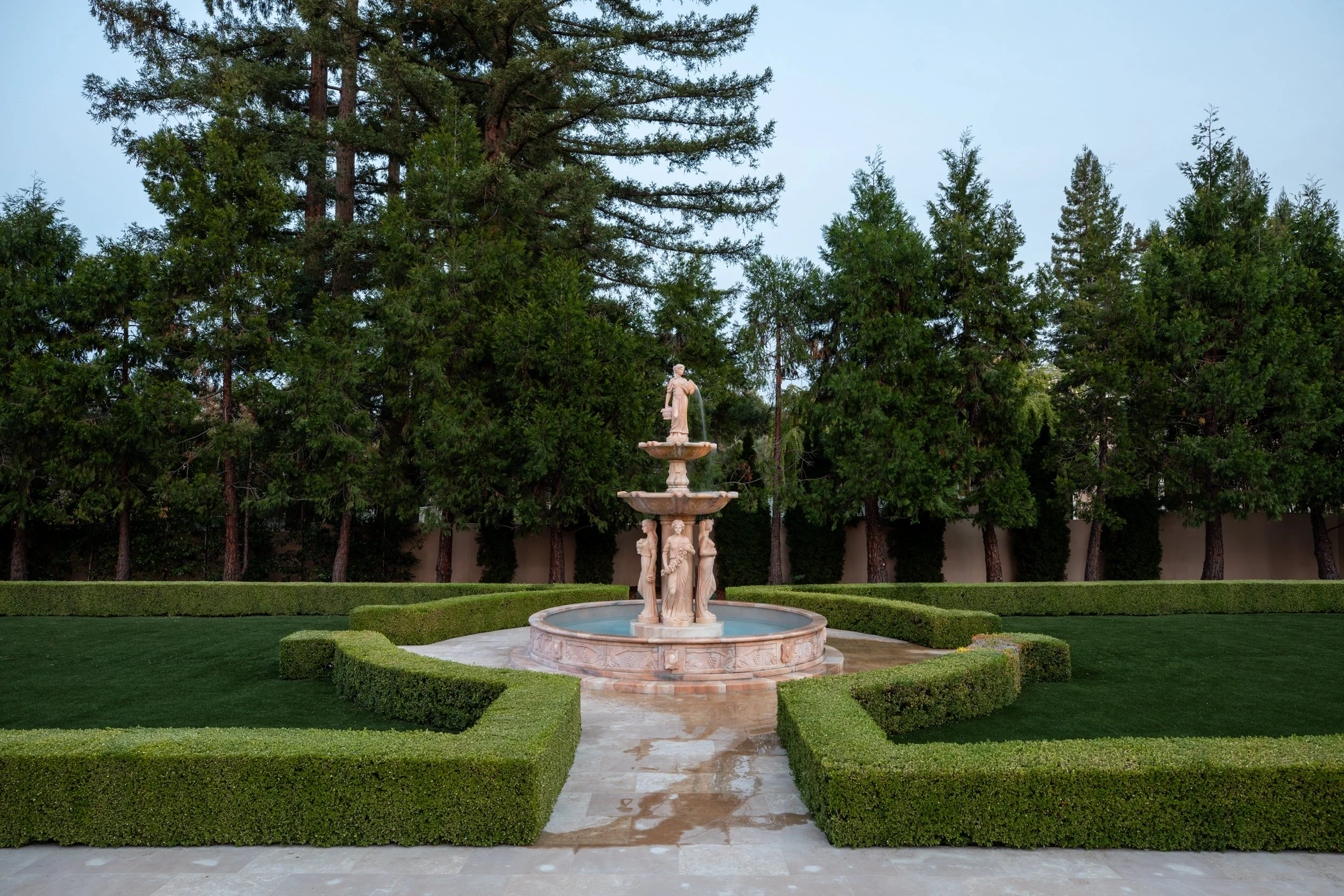 A classical marble fountain with sculptures of women and children, surrounded by neatly trimmed hedges in a garden with tall evergreen trees.