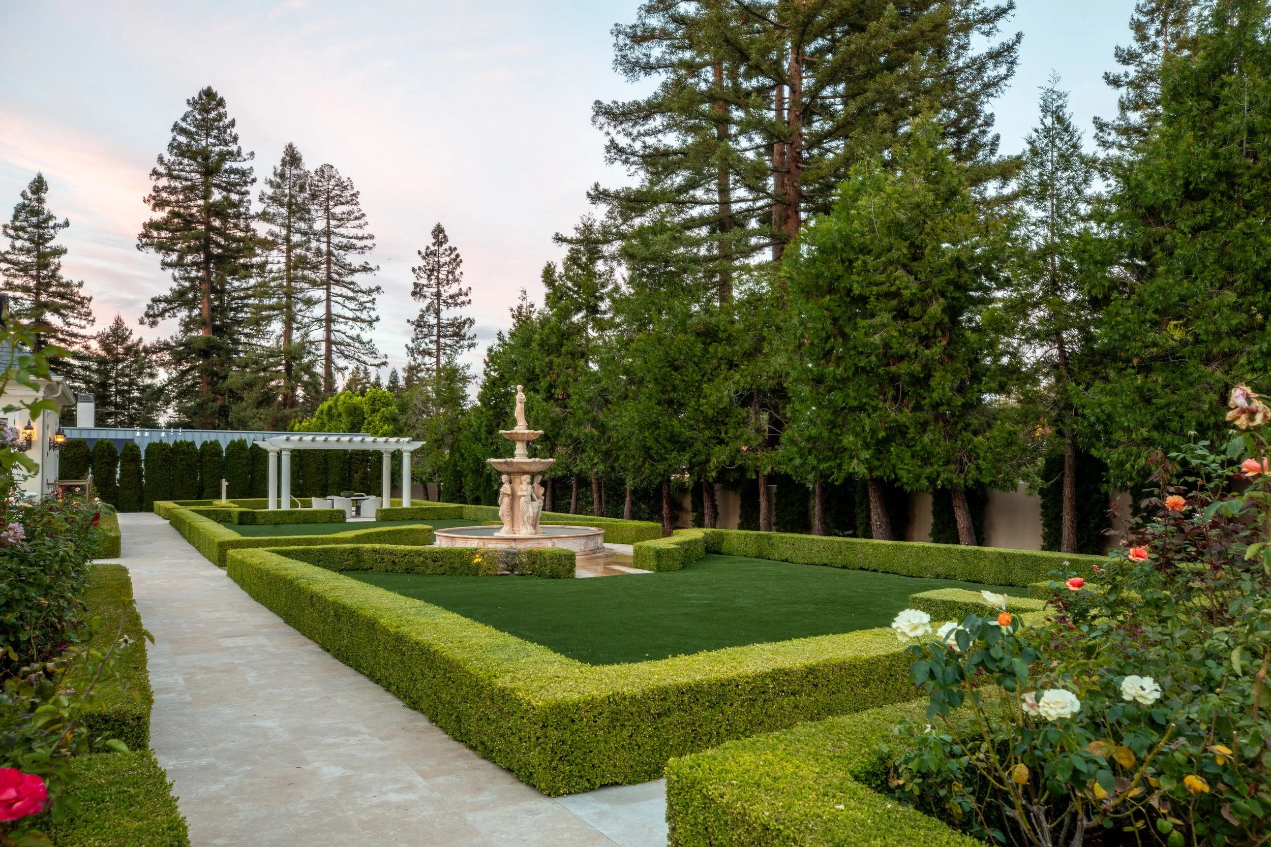 A well-manicured garden with neatly trimmed hedges, a stone pathway, and a classical white fountain featuring statues, surrounded by lush greenery and tall trees in the background.