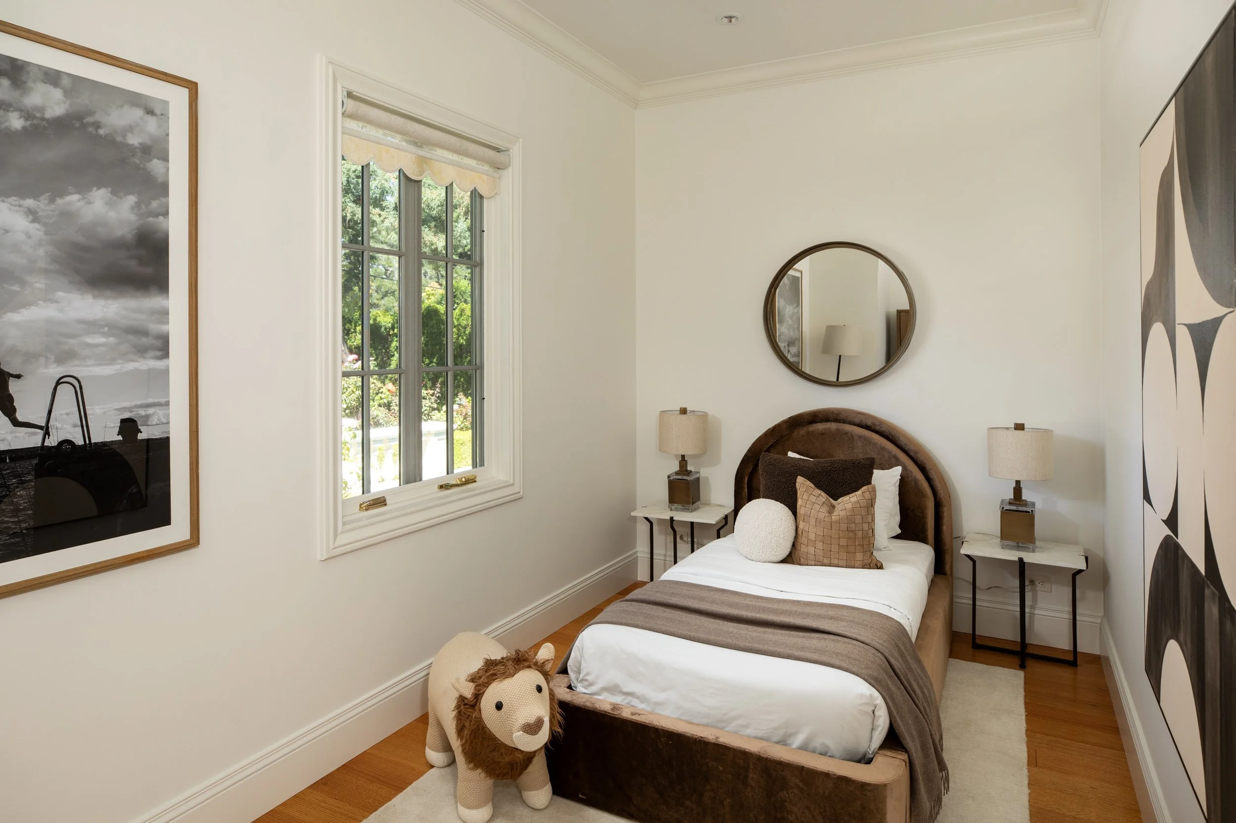 A neatly made bed with brown and white bedding, a plush lion stuffed animal on the floor in front of the bed, two nightstands with lamps, a circular mirror above the bed, a window with a view of trees, and abstract black and white artwork on the wall