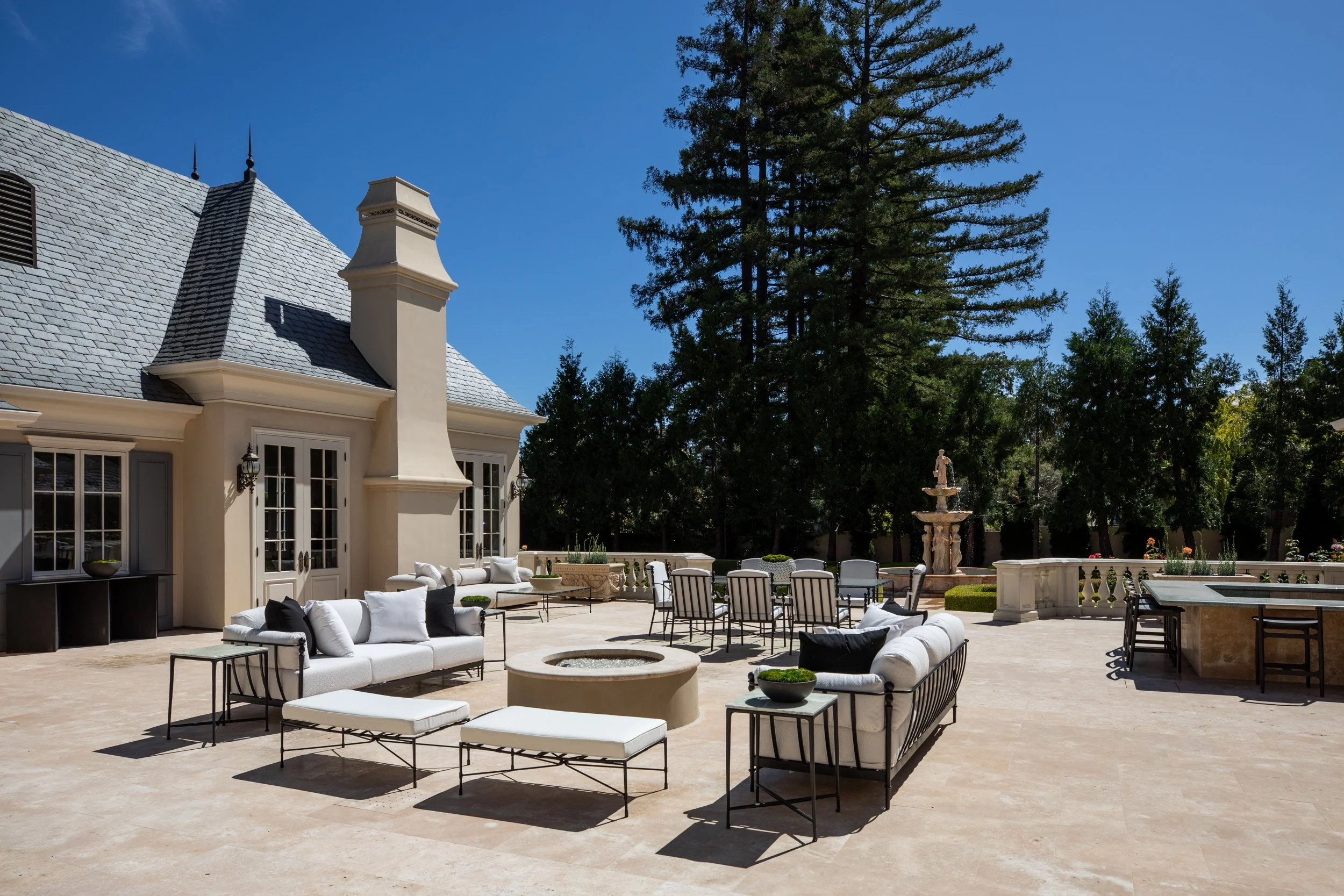 Elegant outdoor patio with white and black modern furniture, a fountain, and lush green trees under a clear blue sky.