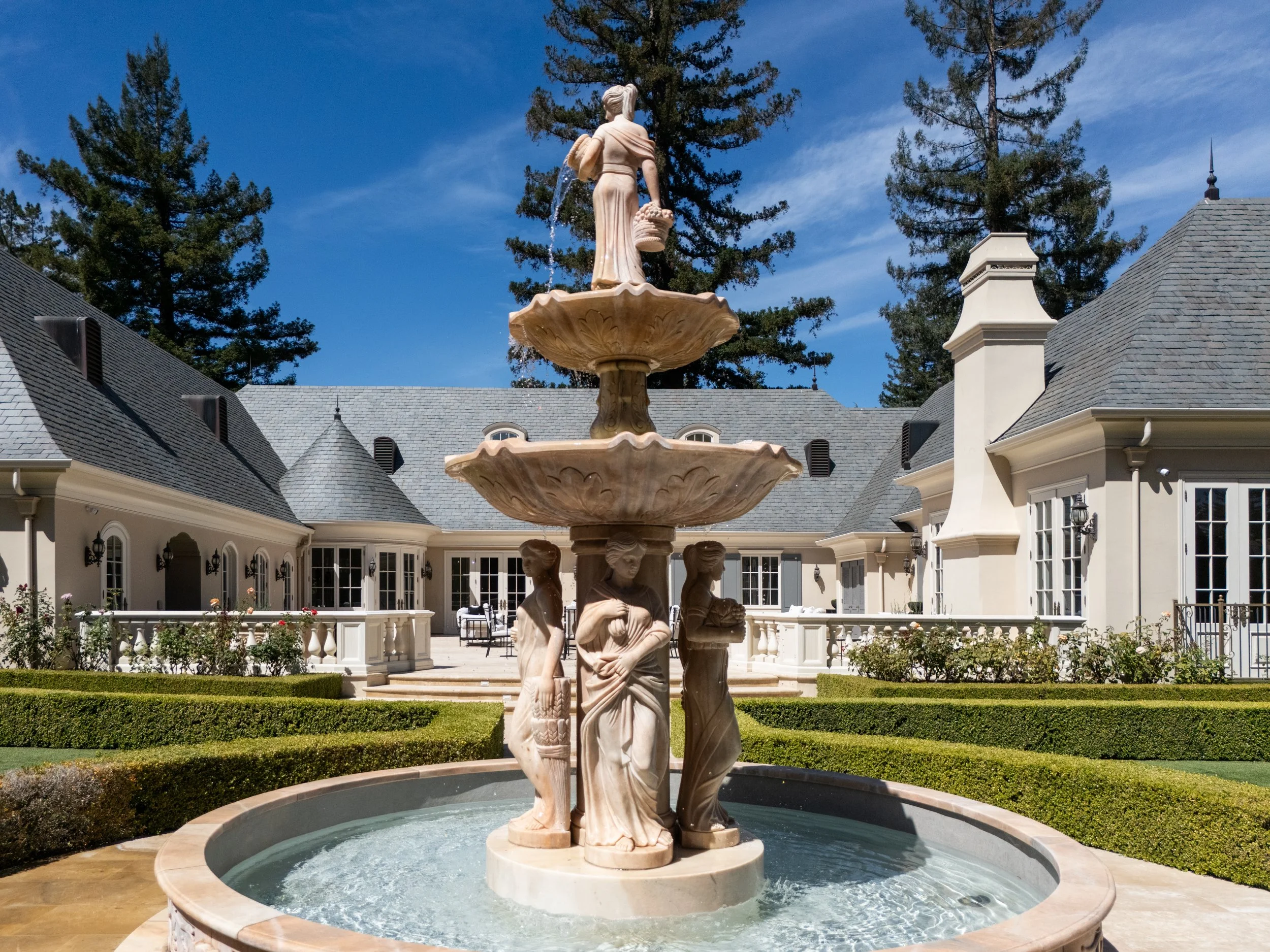 A multi-tiered marble fountain with carved statues of women standing around its base, located in a landscaped courtyard with trimmed hedges and a large mansion-style house in the background, under a clear blue sky with tall pine trees.