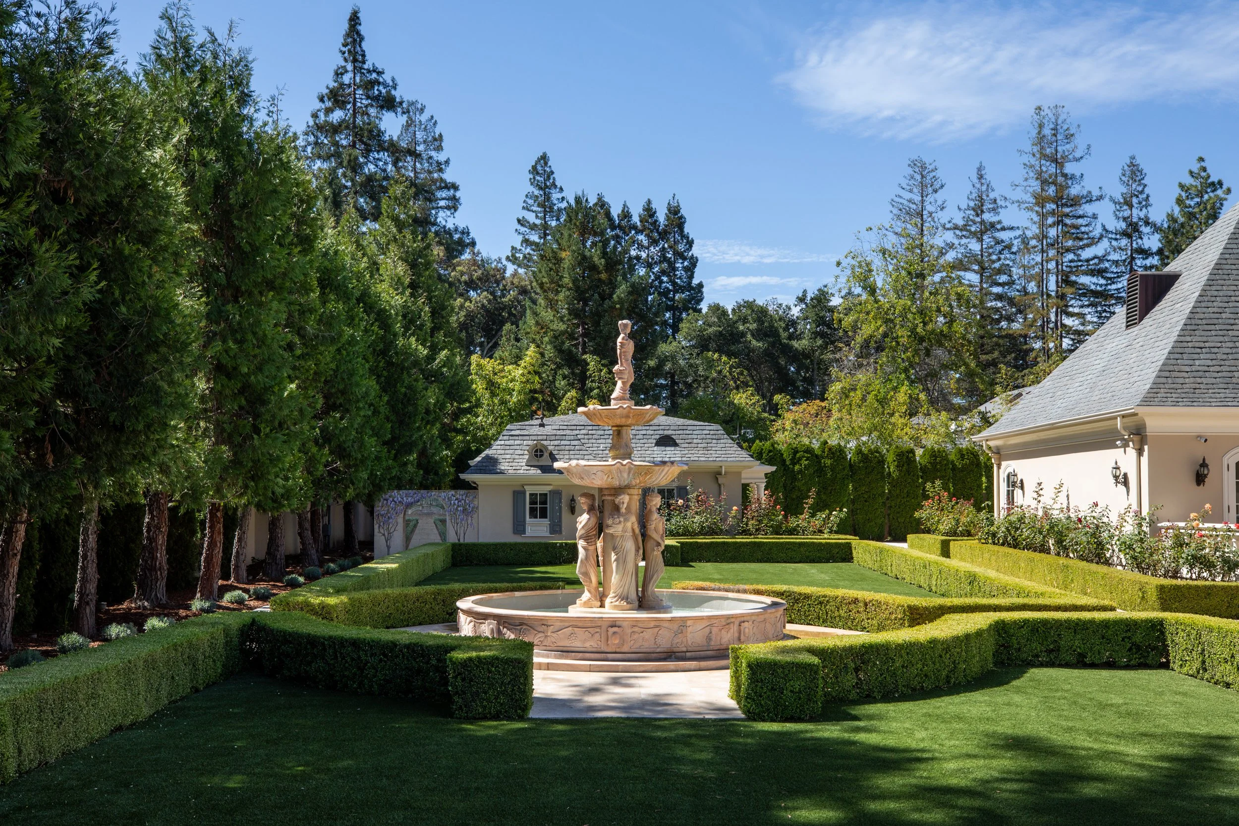 A marble fountain with classical statues in a well-manicured garden surrounded by trimmed hedges, with a house and tall trees in the background under a clear blue sky.
