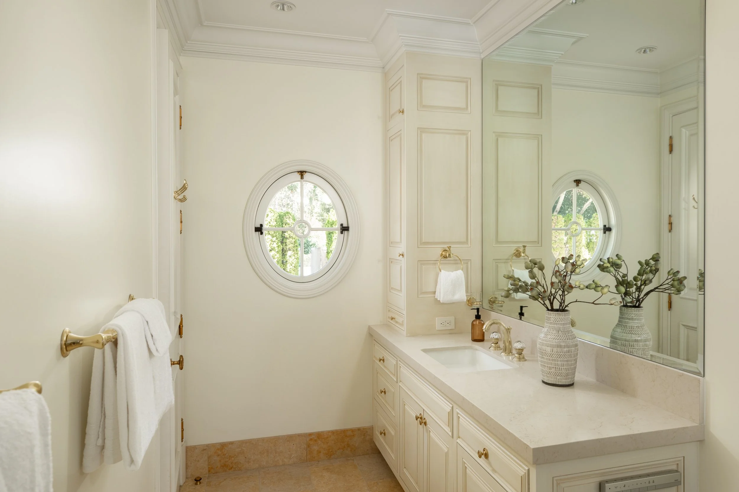 A bright bathroom with cream-colored walls, decorative molding, and two small arched windows with green views outside. The bathroom features a large mirror, a white countertop, a sink, and decorative vases with plants. Gold hardware, including towel 