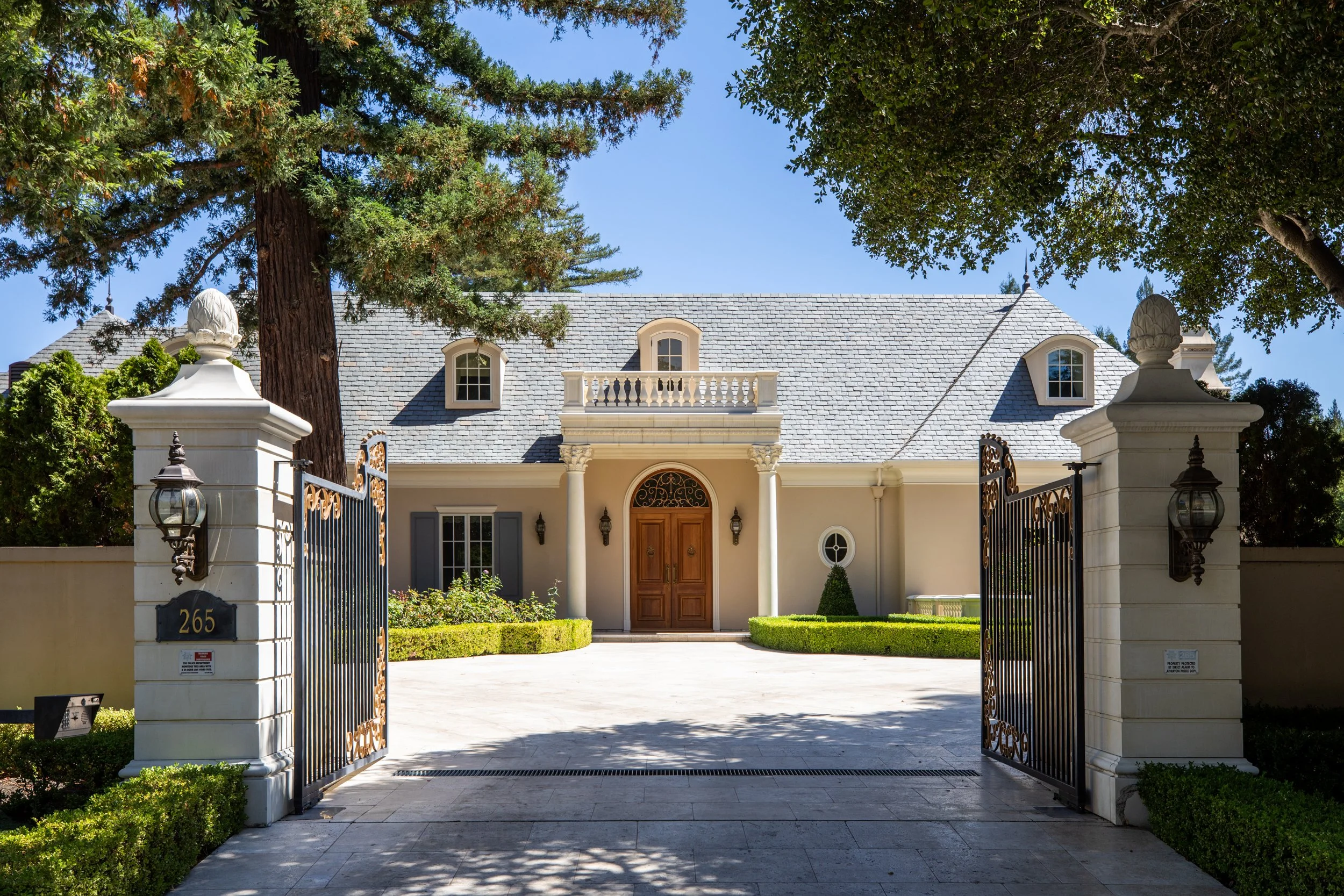 Front view of a large, elegant house with a gray shingle roof, a balcony, and a wooden door, surrounded by trees and a gated fence.