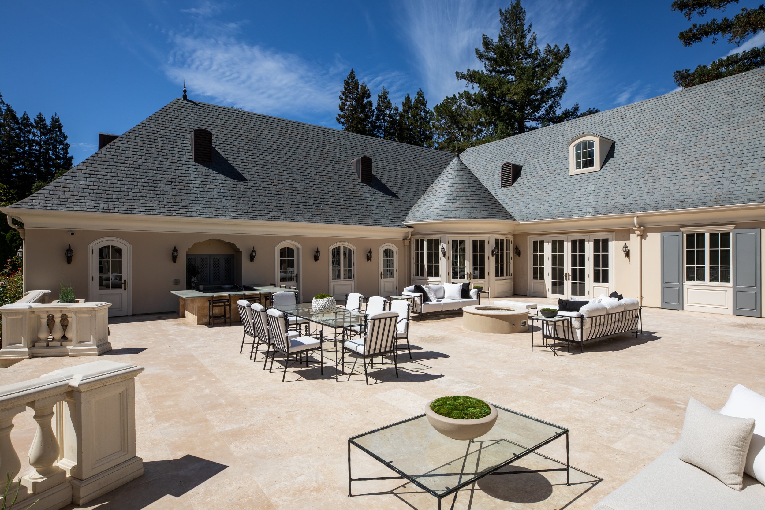 A spacious outdoor patio area with seating and dining furniture in front of a large house with a gray shingle roof, surrounded by tall trees and a partly cloudy blue sky.
