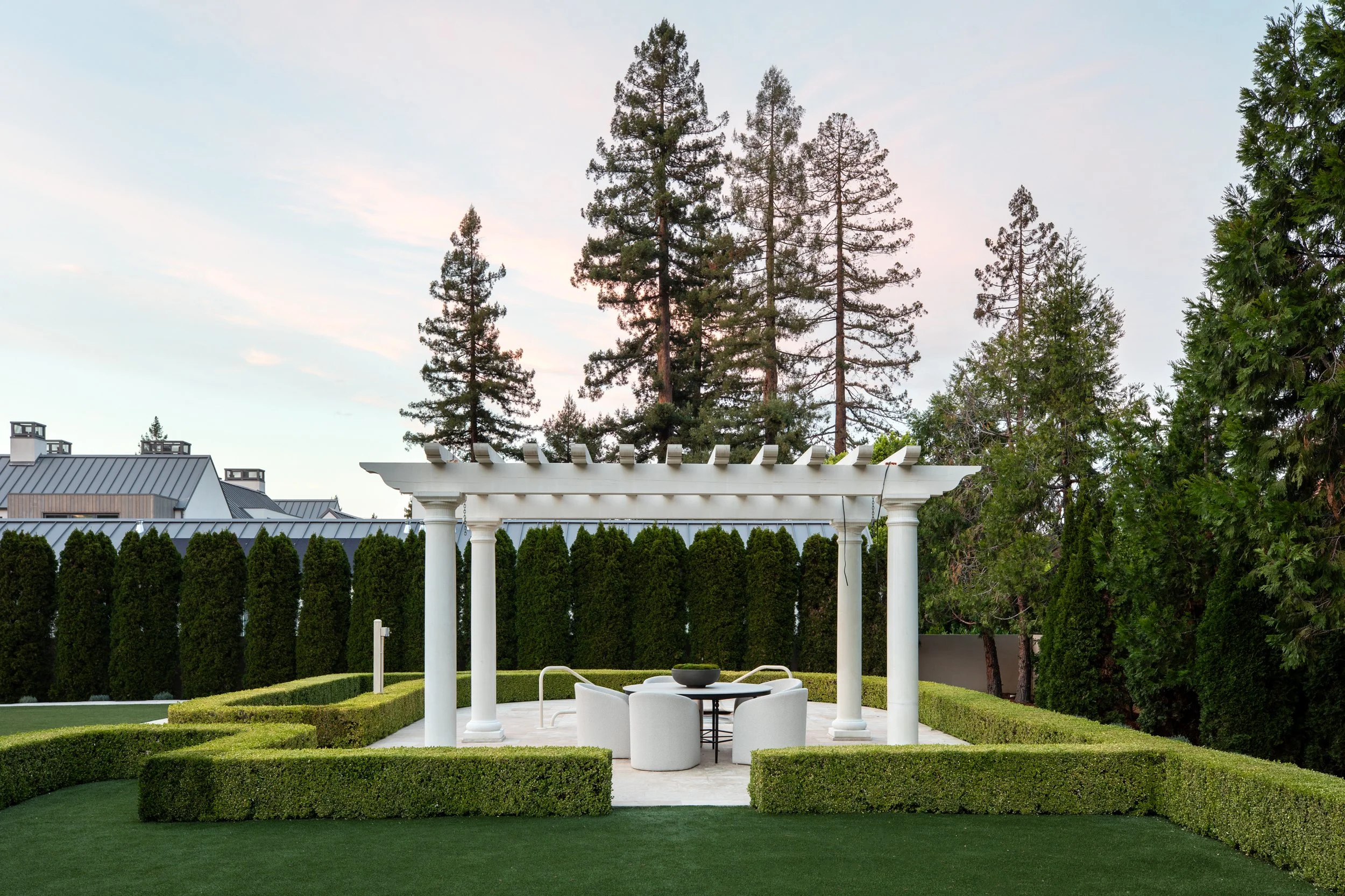 A landscaped backyard with a white pergola, green bushes, and trees in the background under a blue sky.
