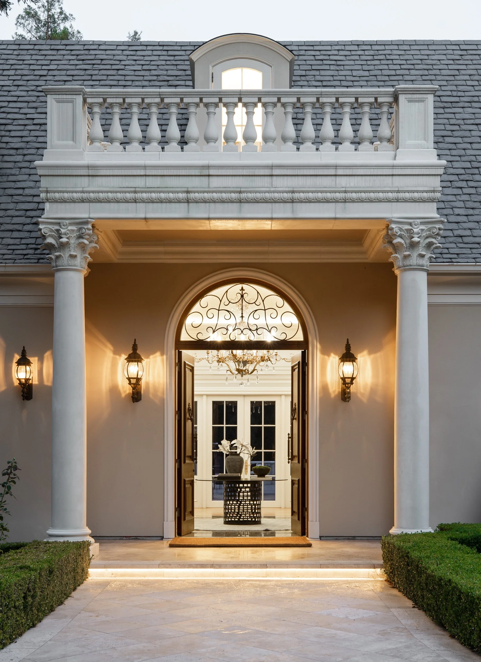 View of a grand entrance with tall white columns and a large arched doorway with open wooden doors, leading into a luxurious interior with a chandelier and decorative windows.
