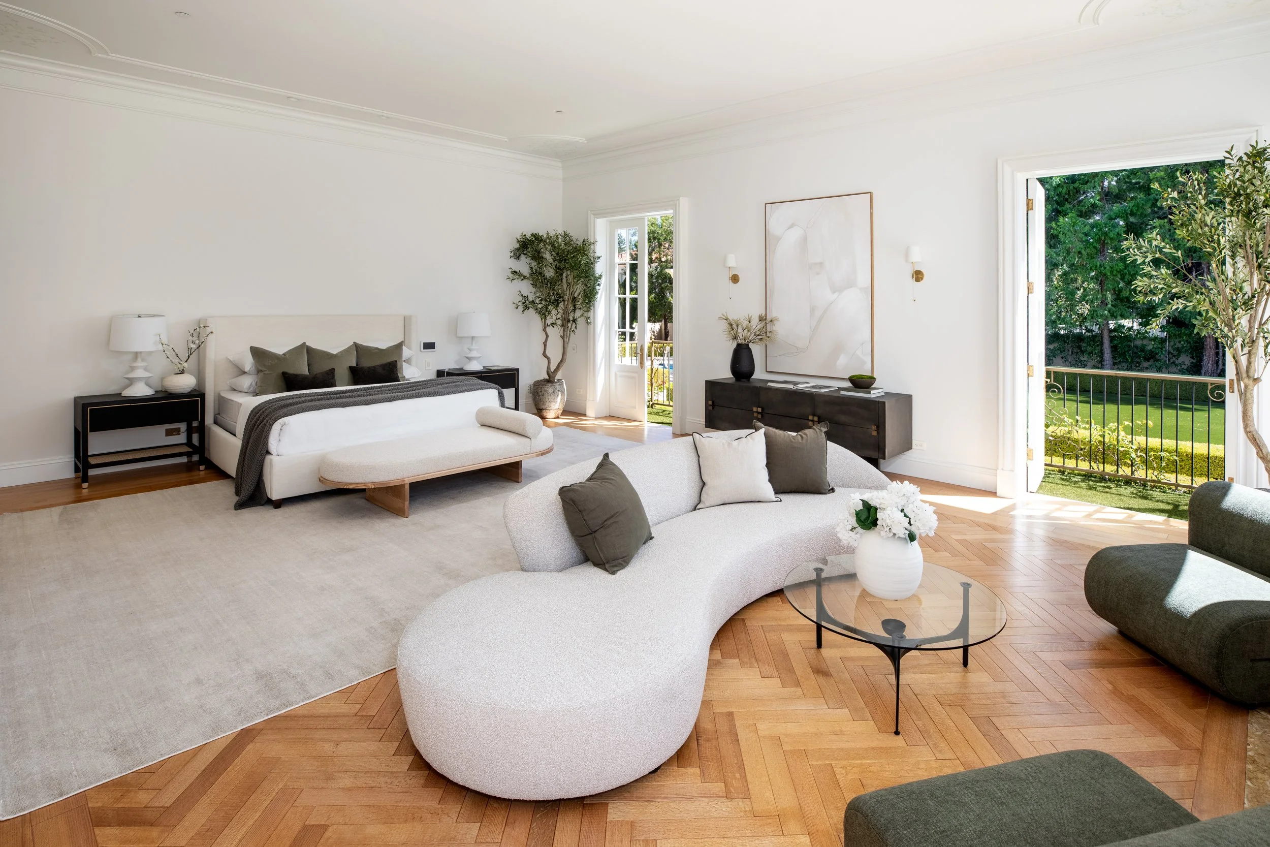 Living room with white walls, large windows, a white and beige sofa, a curved armrest, and a round glass coffee table. There are decorative cushions on the sofas, a black side table with white lamps, a large potted plant, a black console table with a