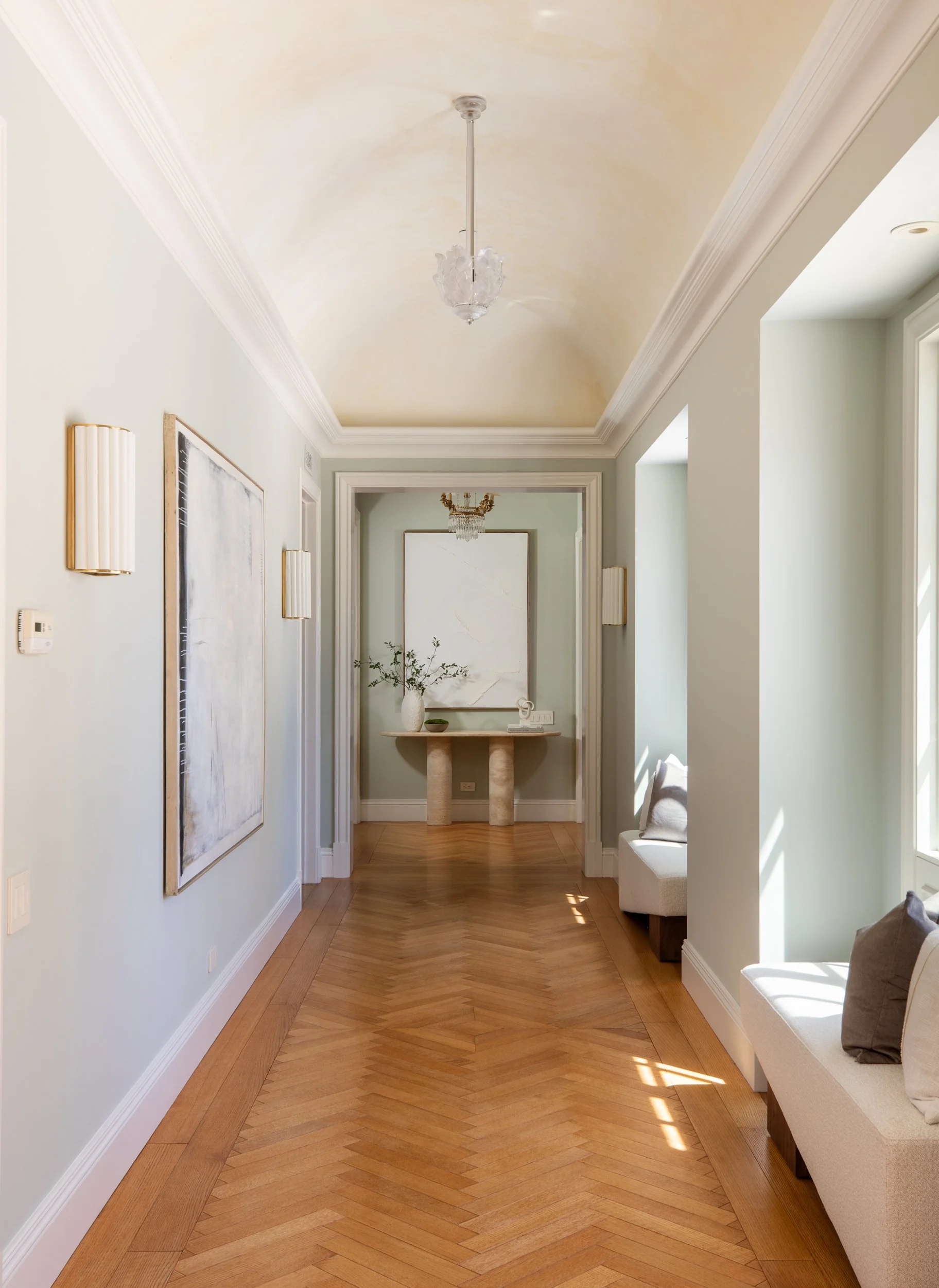 An elegant hallway with a vaulted ceiling, wooden flooring, wall-mounted lights, a piece of artwork, a console table with decorative items, and a window seat with cushions.