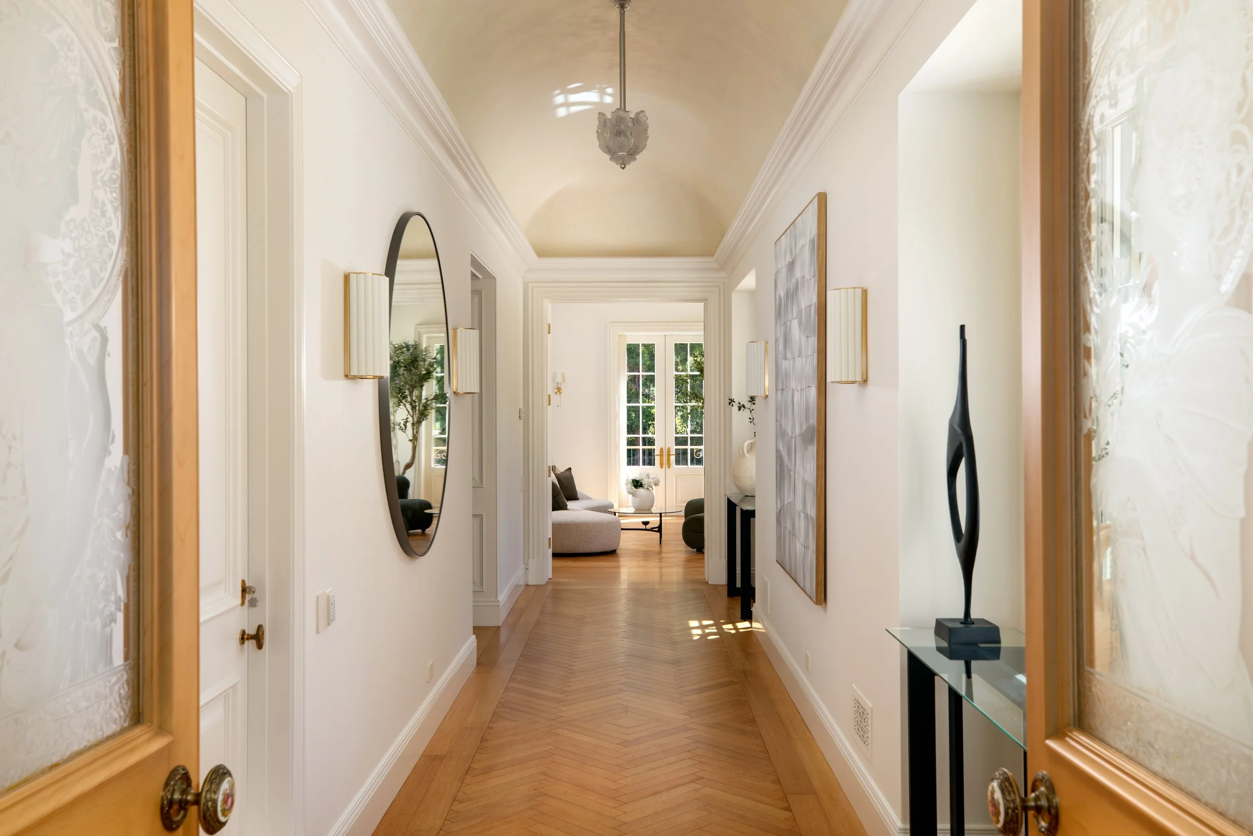 A hallway view into a bright living room with large windows, wooden floors, and furniture.