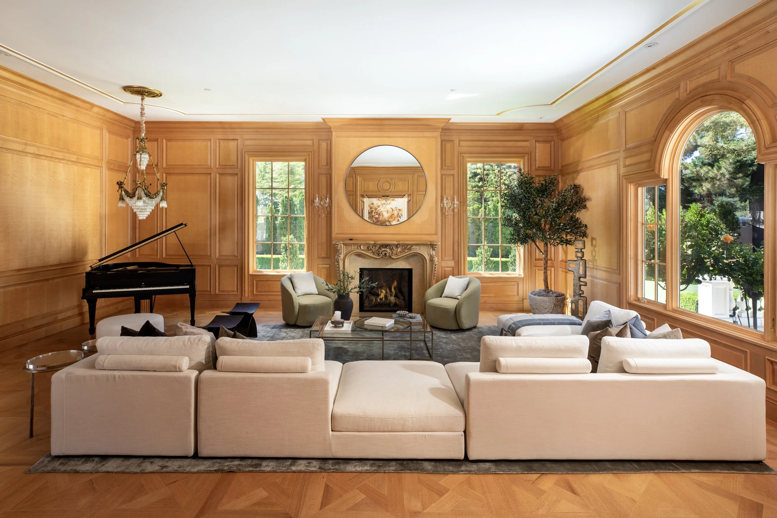 Elegant living room with wood-paneled walls, large windows, a grand chandelier, a white sofa, armchairs, a grand piano, a fireplace with a mirror above, indoor plants, and a coffee table.