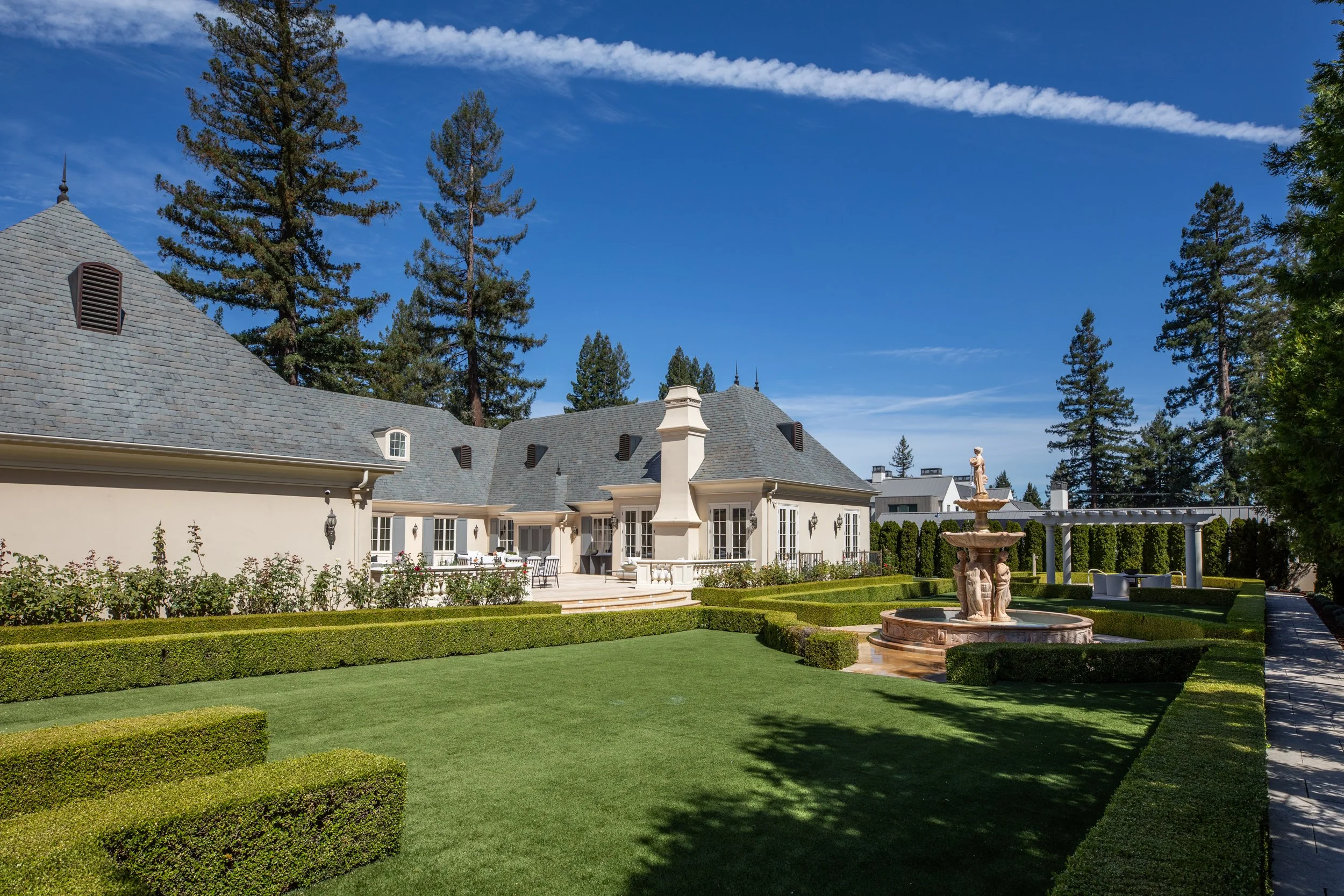 A large, elegant house with a gray slate roof, surrounded by a well-maintained garden with trimmed hedges, a fountain with statues, and tall pine trees under a partly cloudy blue sky.