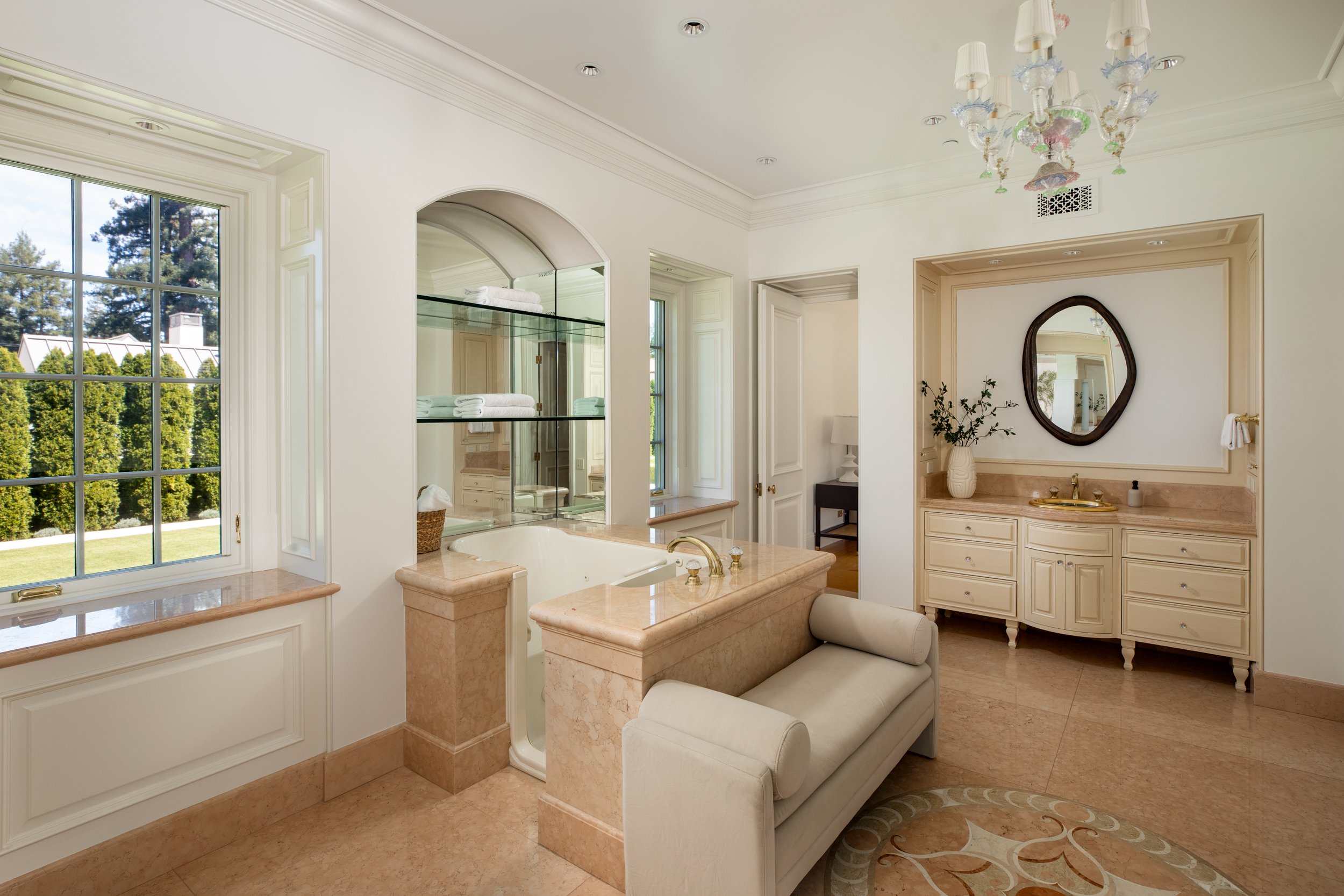 Elegant bathroom with large window, marble countertops, a bathtub, a vanity with a mirror, and decorative chandeliers.