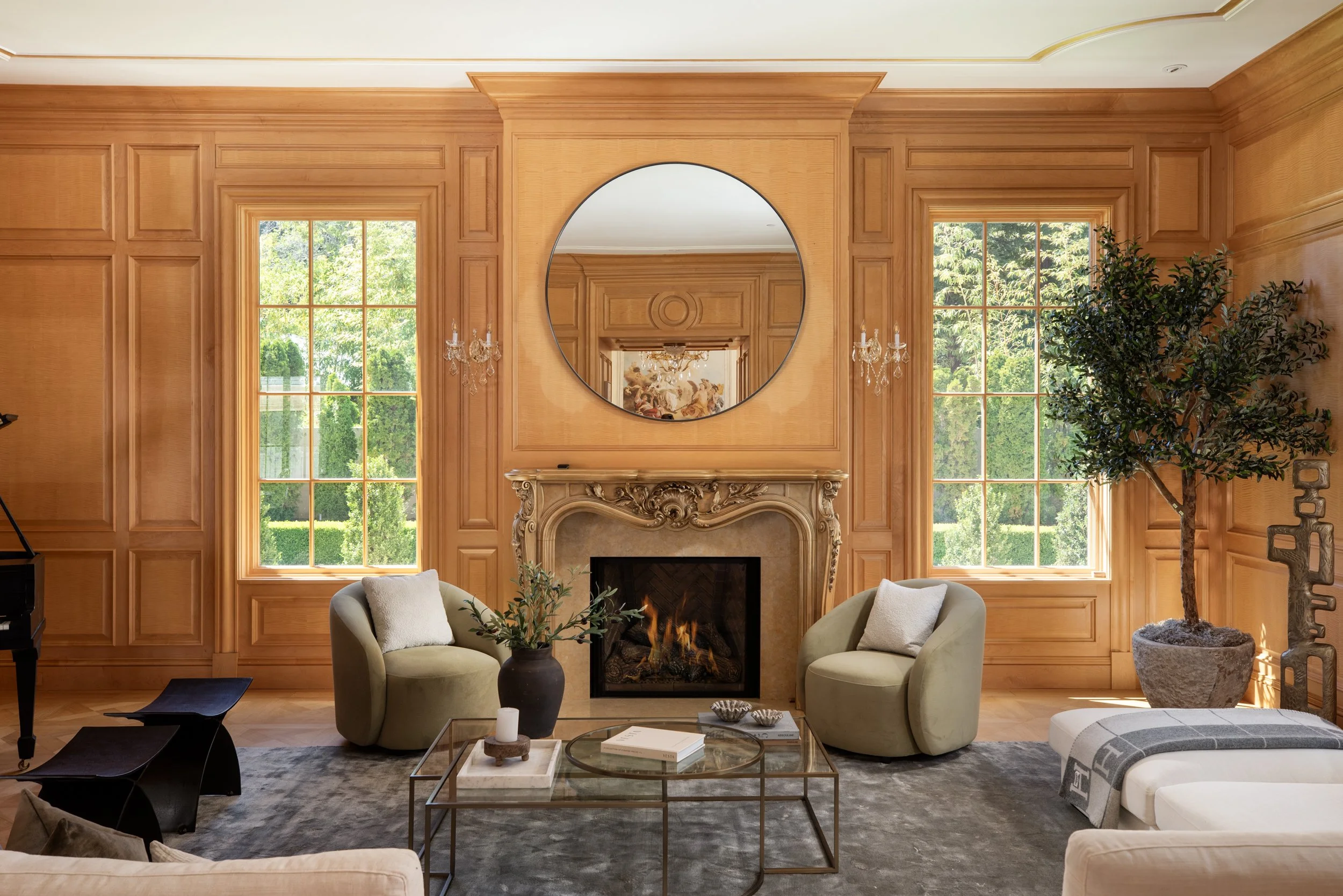 Elegant living room with wooden wall paneling, large windows showing a green outdoor scene, a fireplace, a round mirror above the fireplace, two armchairs, a glass coffee table with decorative items, chandeliers, and a tall potted plant.