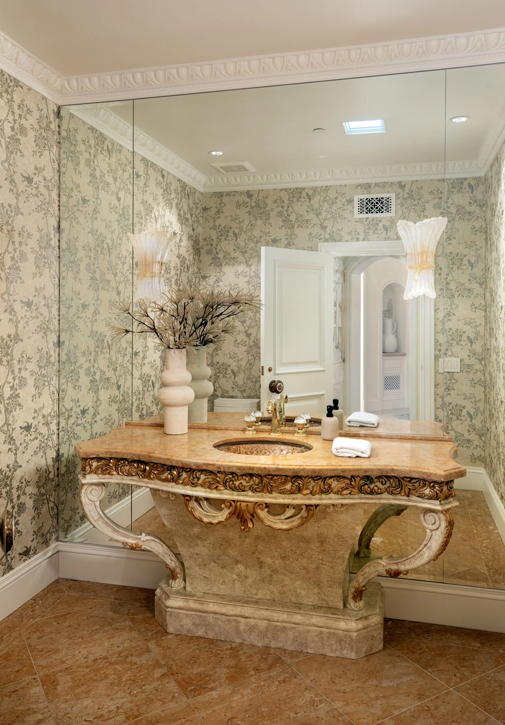 A bathroom with a large mirror, ornate wooden vanity with a marble countertop, decorative vases with dried branches, and a framed mirror with a floral wallpaper background.