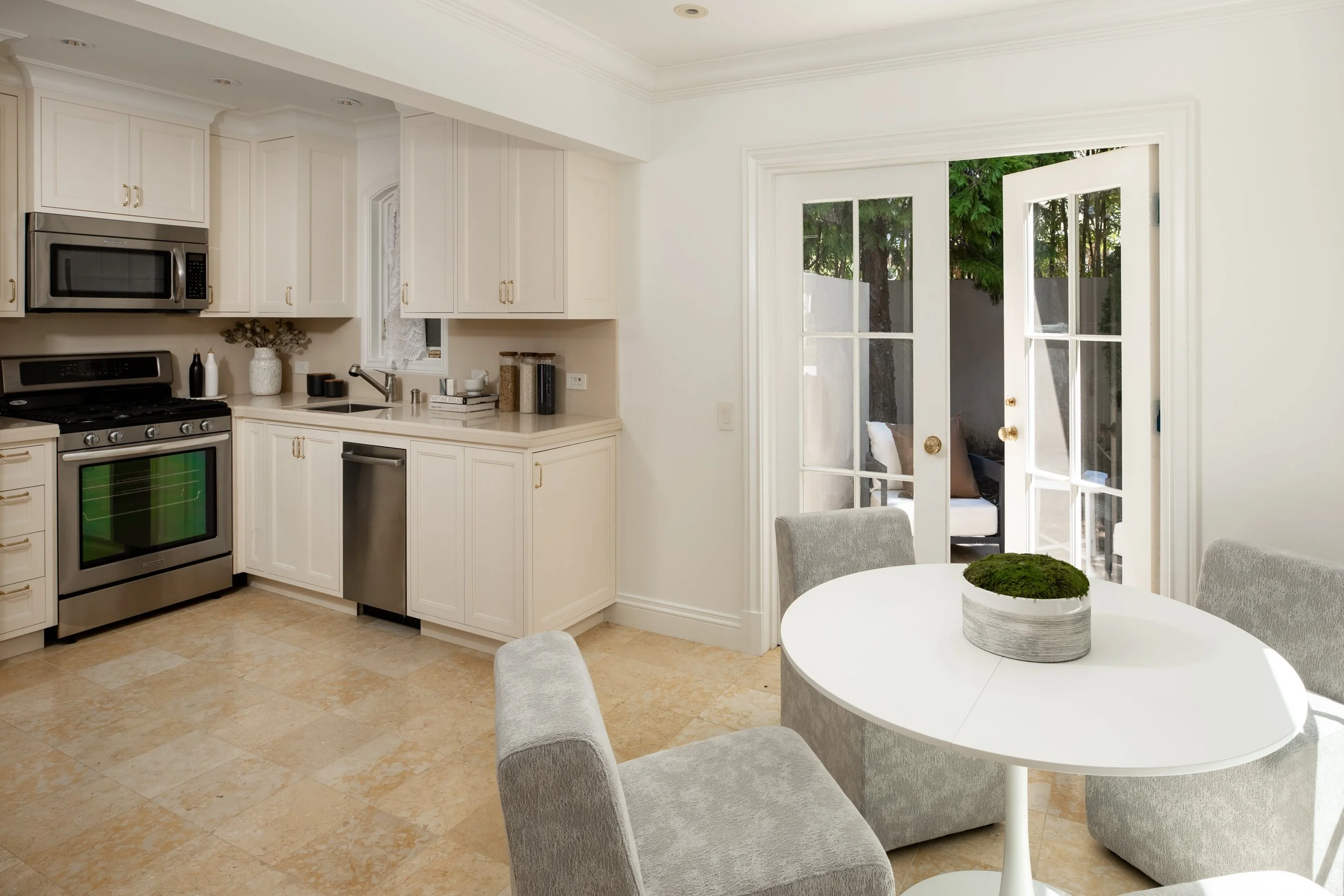 A bright kitchen and dining area with white cabinets, stainless steel appliances, a round white table with four gray upholstered chairs, and a view through glass-paned French doors to an outdoor patio and greenery.