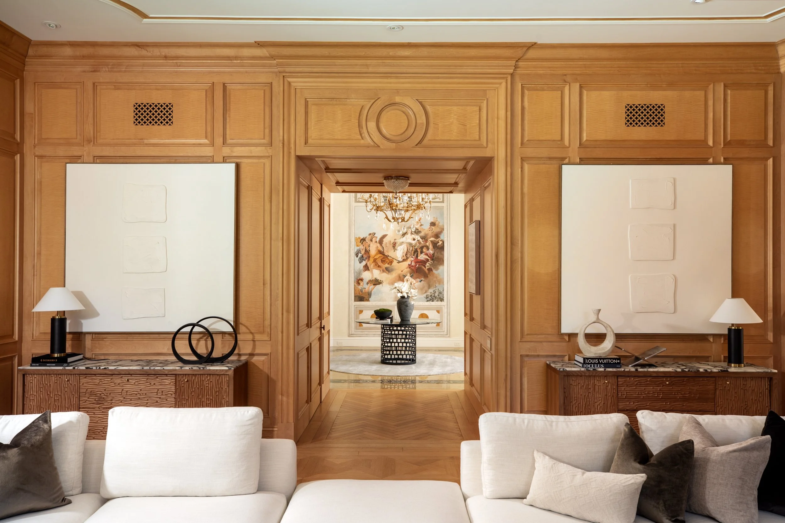 A luxurious living room with wooden paneling and a doorway leading to another room with a chandelier and large mural painting of angels and humans.