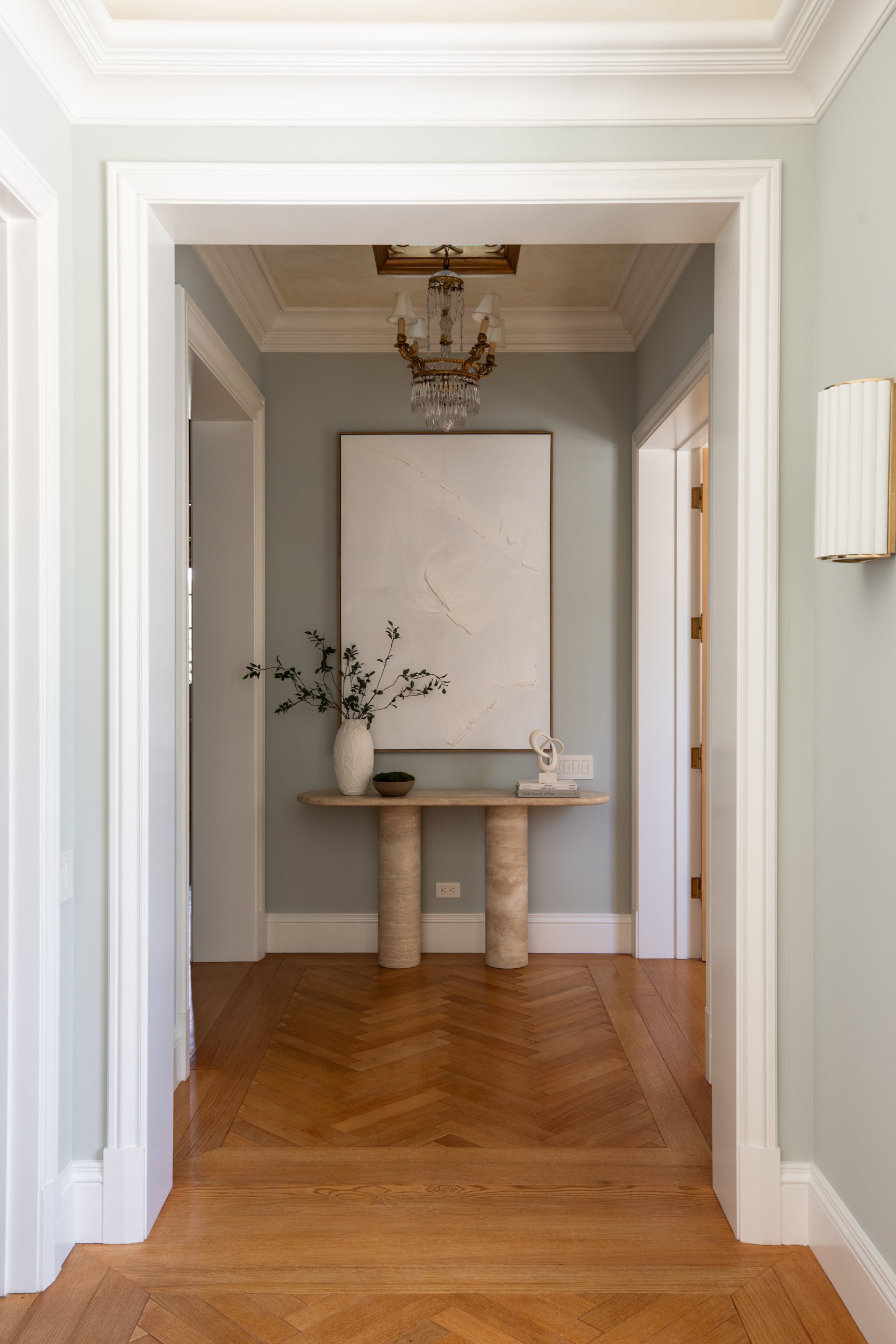 A hallway with light-colored walls, white door frames, a wooden herringbone floor, and a decorative chandelier hanging from a rectangular ceiling cut-out. A console table with a large white vase with green branches, a small bowl, and a sculpture is a