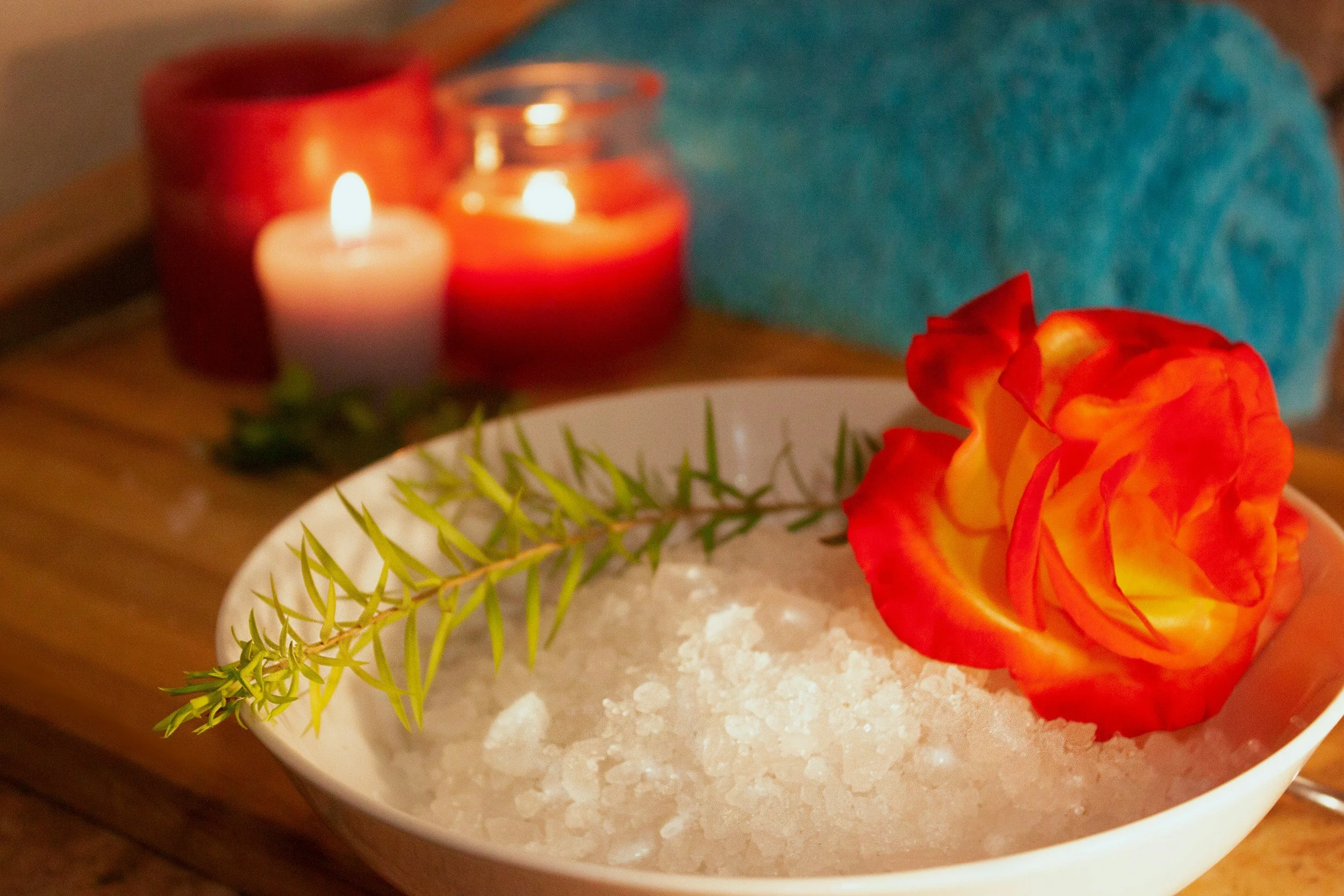 A bowl filled with white bath salts, garnished with a sprig of greenery, a bright orange and yellow rose, and surrounded by lit candles in red and orange holders, creating a relaxing spa setting.
