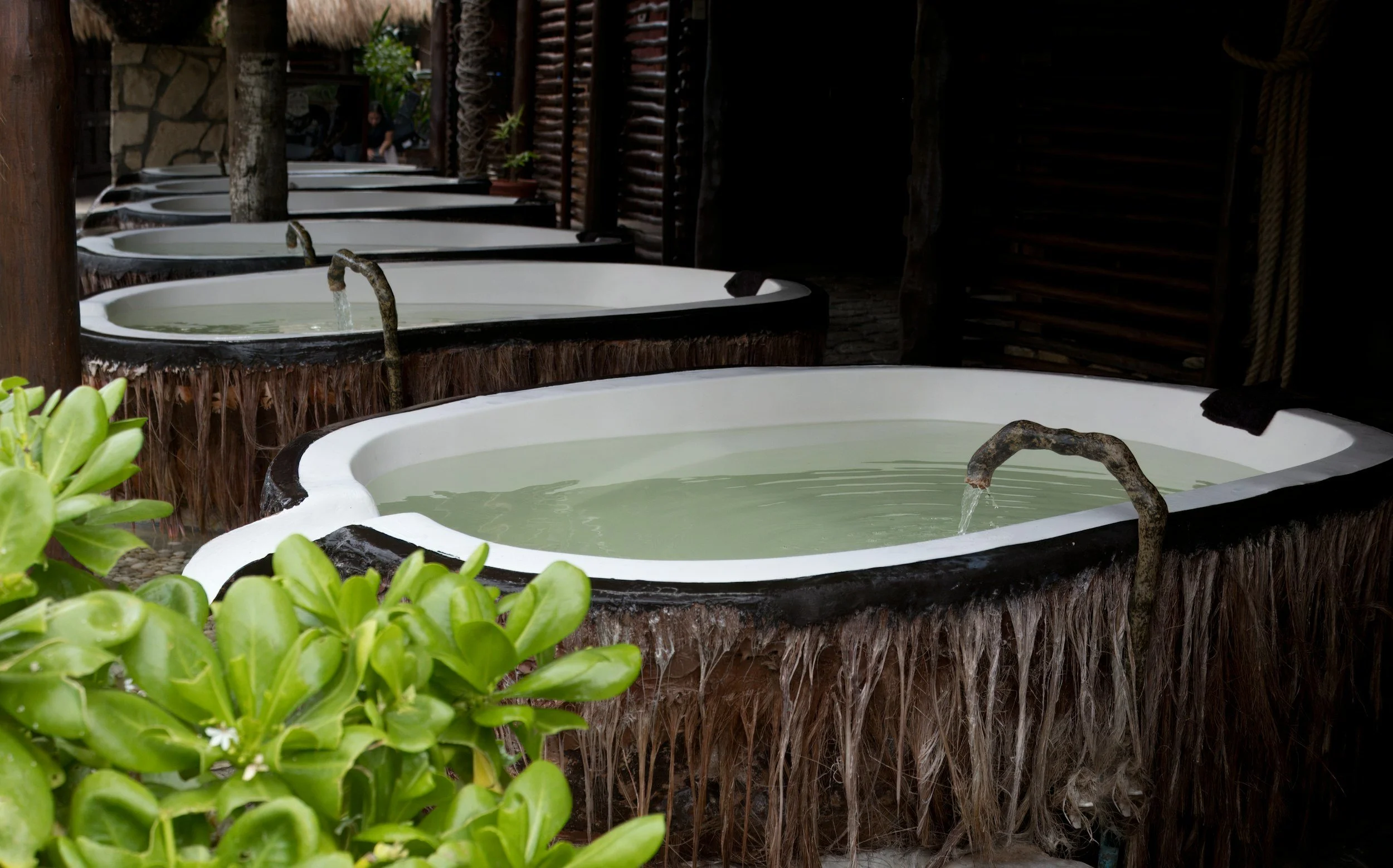 Row of rustic outdoor bathtubs with water spouts, surrounded by tropical greenery and wooden structures.