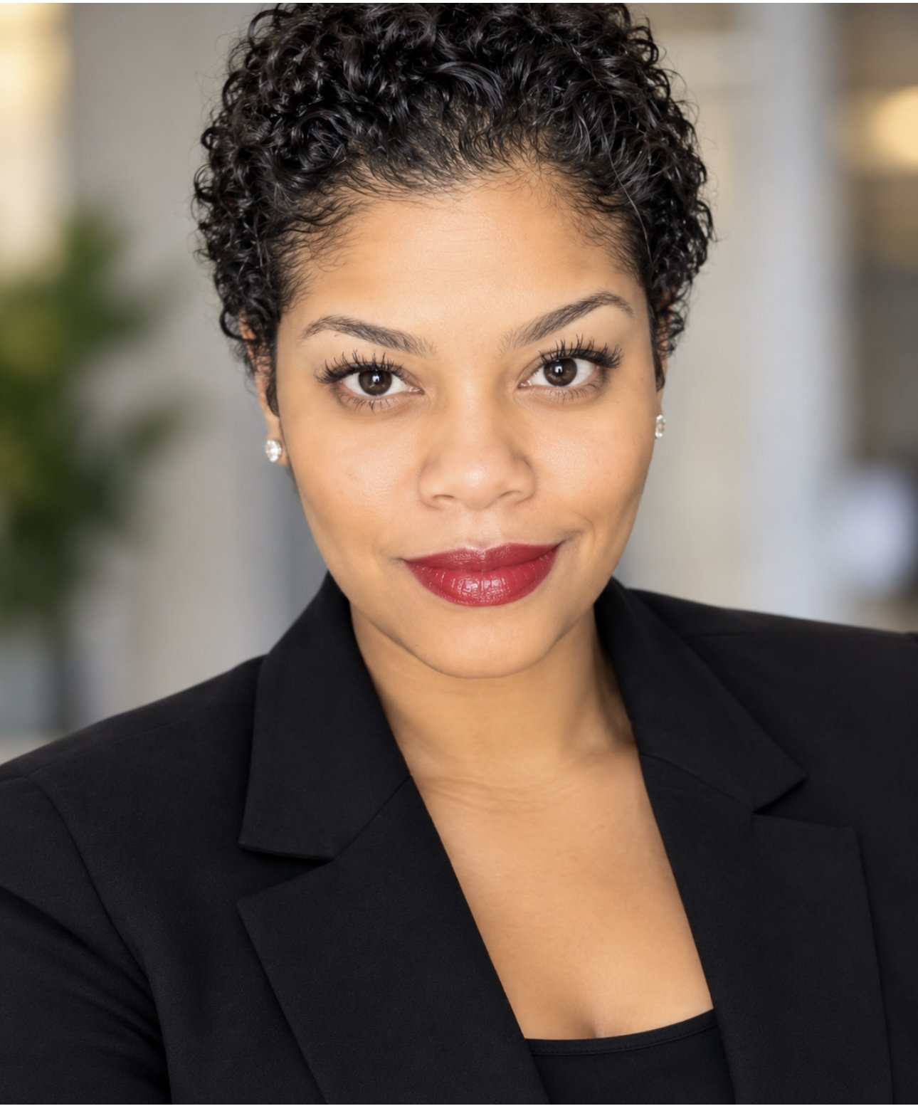 Professional woman with short curly black hair, wearing a black blazer and red lipstick, smiling in an indoor setting.