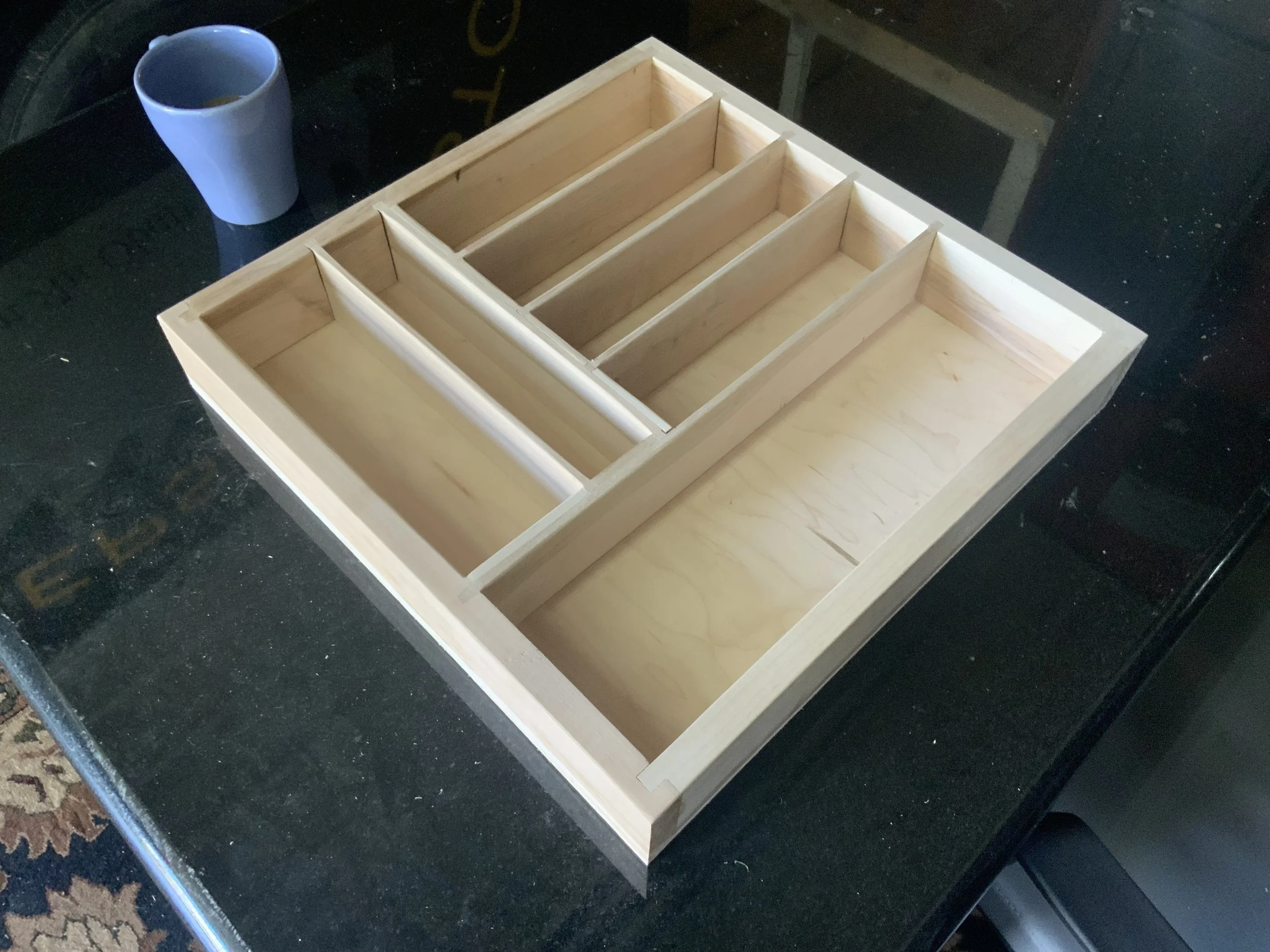 Unfinished wooden box with multiple compartments on a black surface, a white cup on the left side.