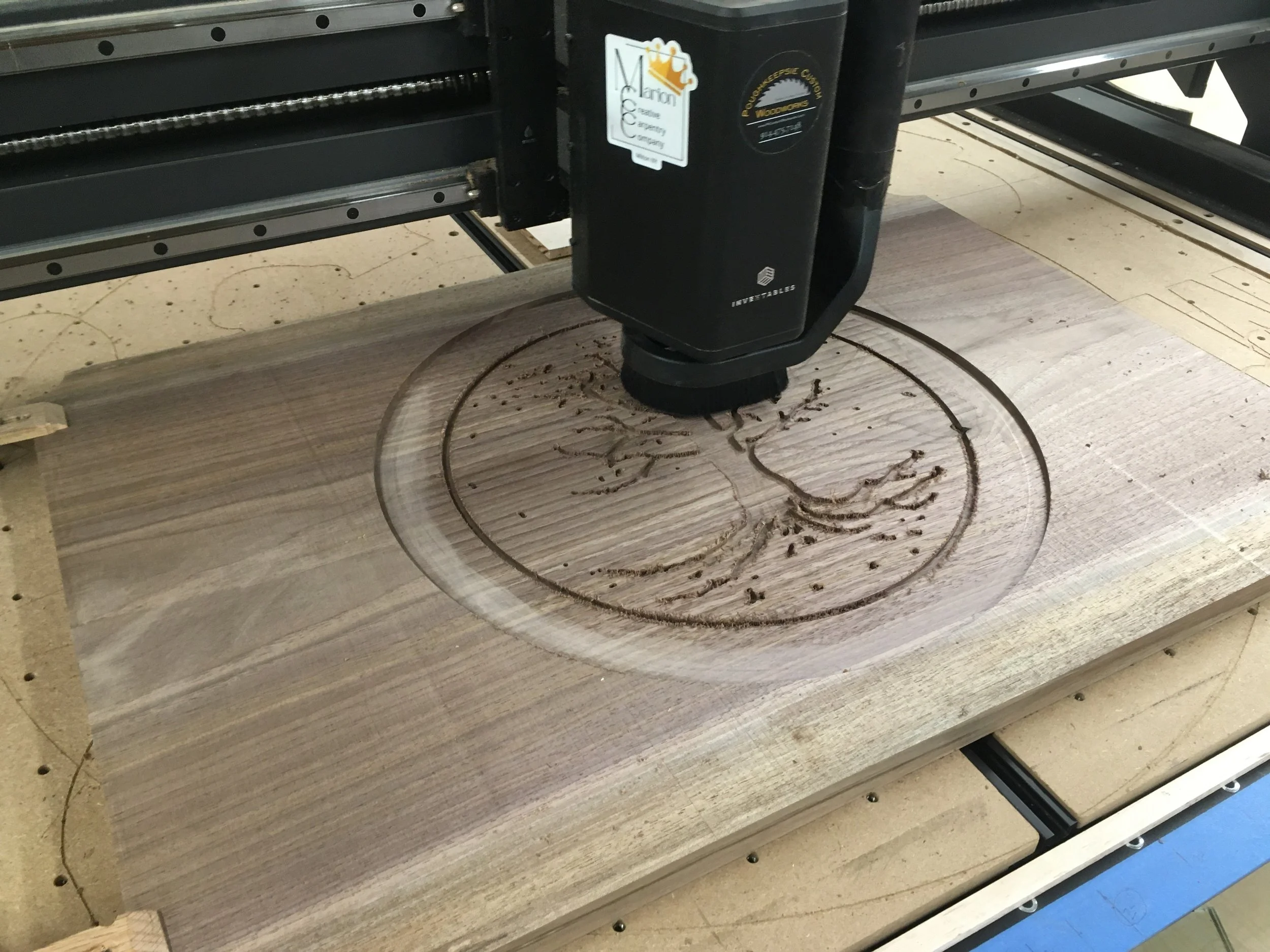 A CNC machine carving a design into a piece of wood. The wood is secured on a flat surface, and sawdust is visible around the carving area.