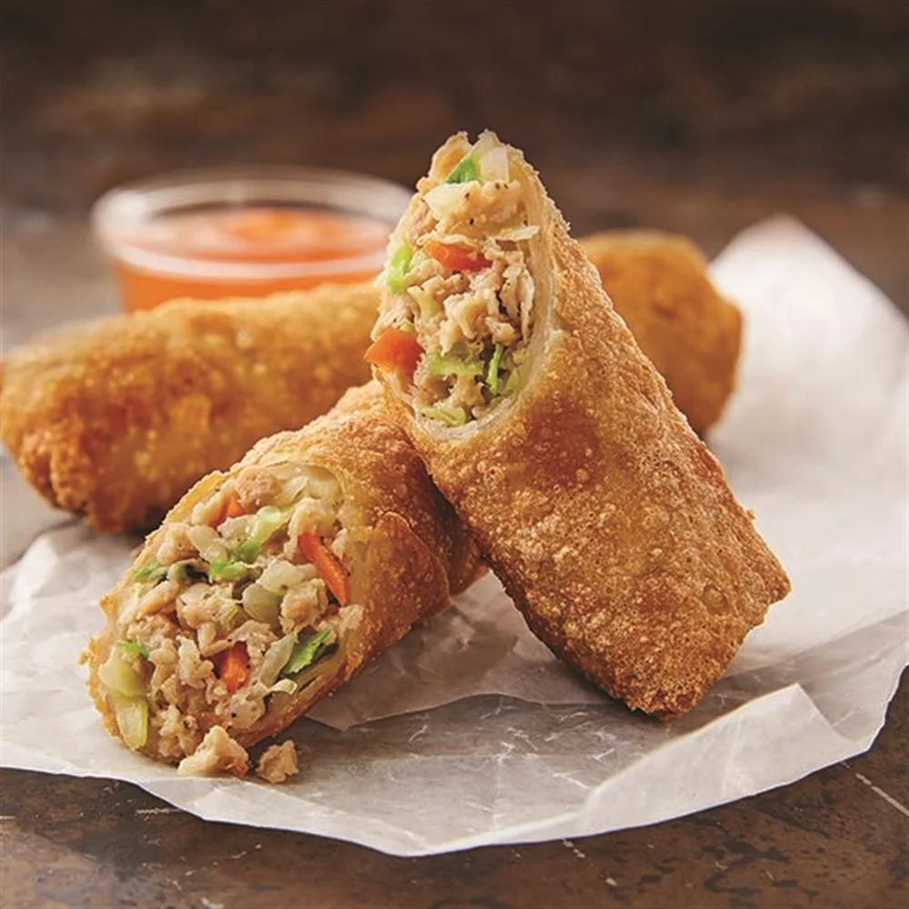 Two fried egg rolls filled with vegetables and chicken, one is cut in half to show the filling, with a side of dipping sauce in a small glass cup in the background.
