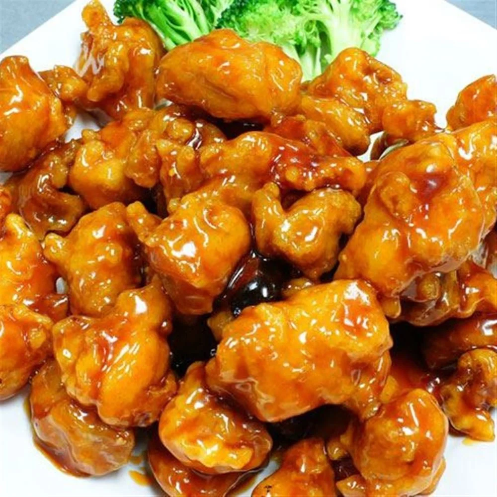 A plate of sweet and sour chicken with chunks of crispy chicken coated in an orange sauce, garnished with some green broccoli and lettuce.