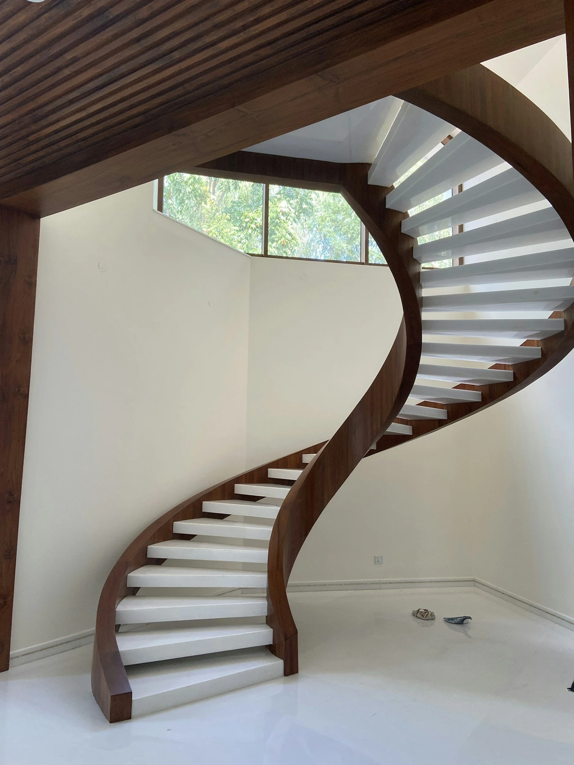 Photo of a spiral staircase with wooden handrail and white steps, located inside a building with large windows and a view of trees outside.