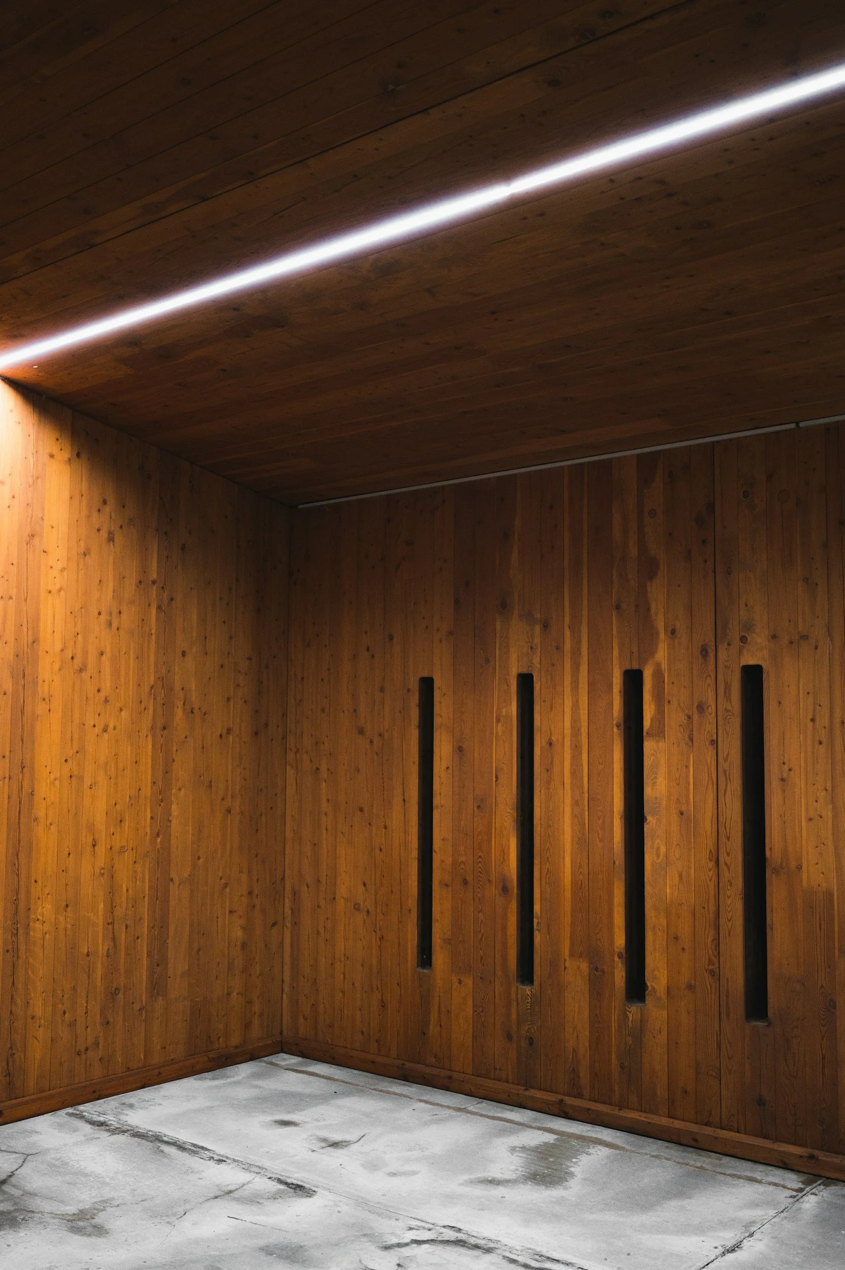 Interior space with wooden paneled walls and ceiling, featuring four vertical black openings on the wall, and a concrete floor with visible cracks and stains.