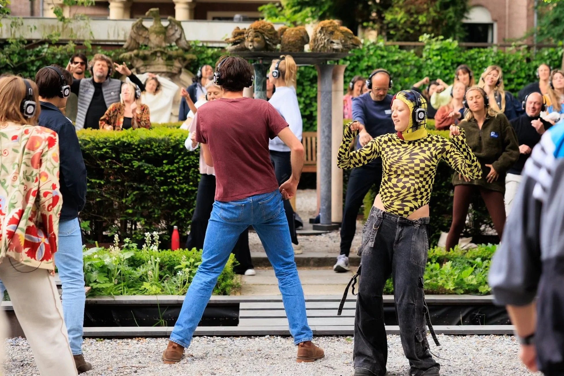 A woman in a yellow and black checkered bodysuit poses with flexed arms while a group of people, some wearing headphones, dance and enjoy a music event outdoors in a garden setting.