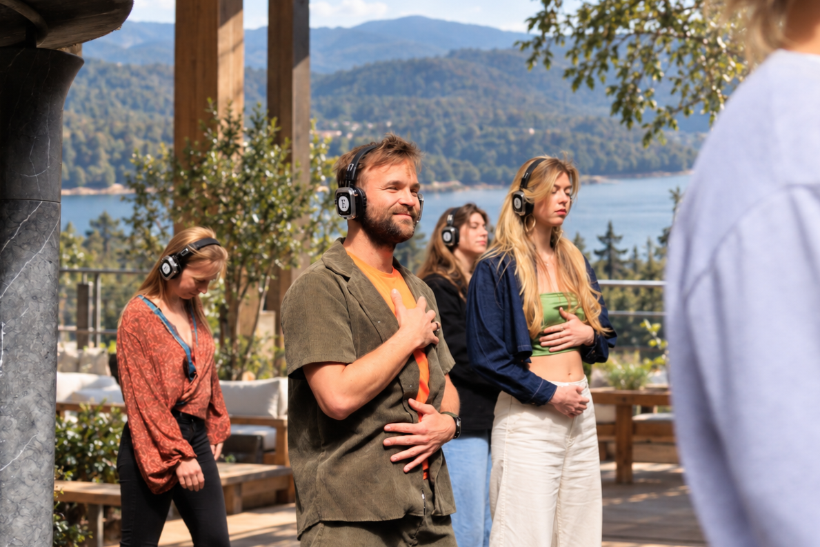 Group of people standing outdoors, wearing headphones, with a scenic lake and mountains in the background, some with eyes closed and hands on their chests.