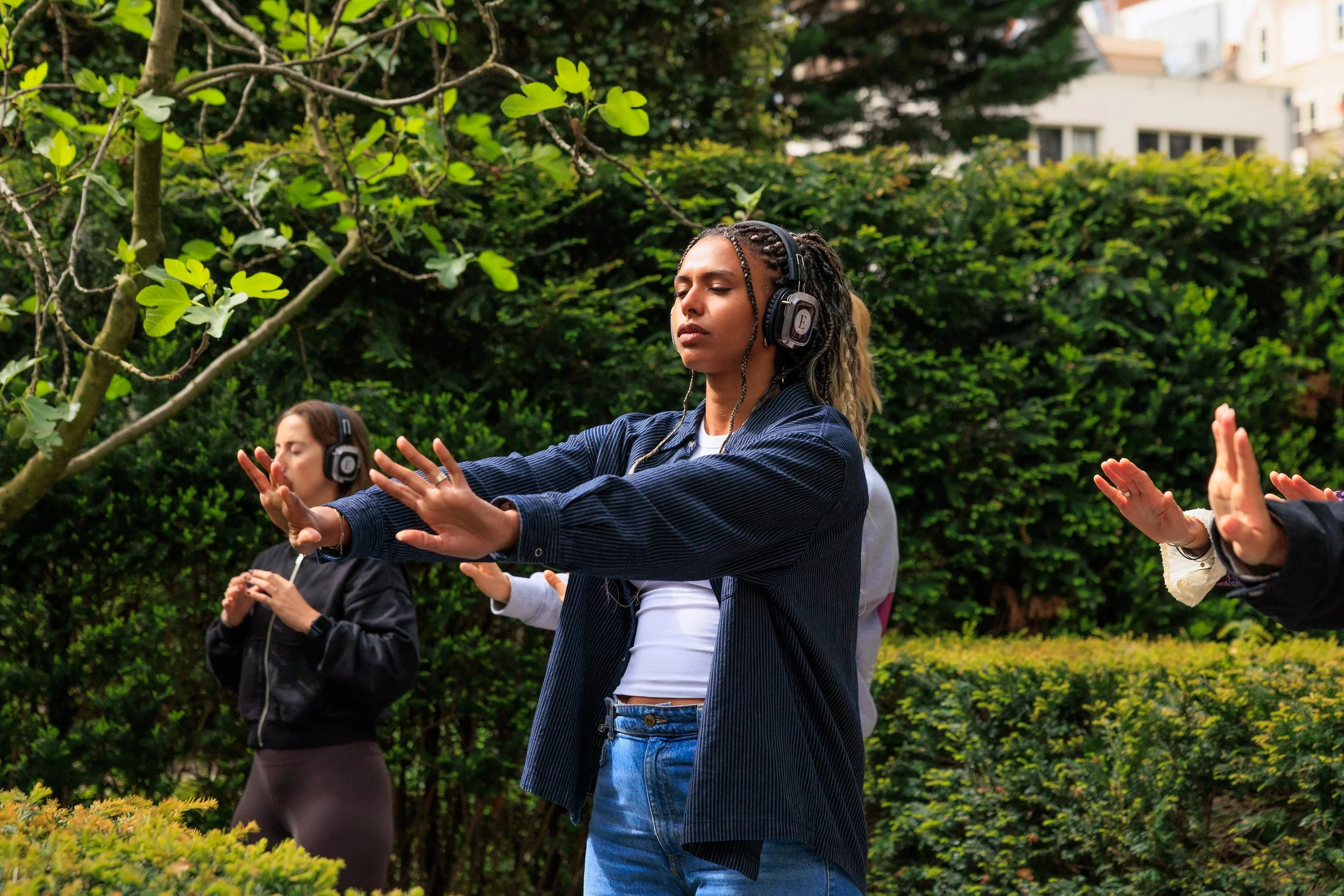 A young woman with braids, wearing headphones, practicing meditation or yoga outdoors with eyes closed and hands raised in a calming gesture, surrounded by greenery.